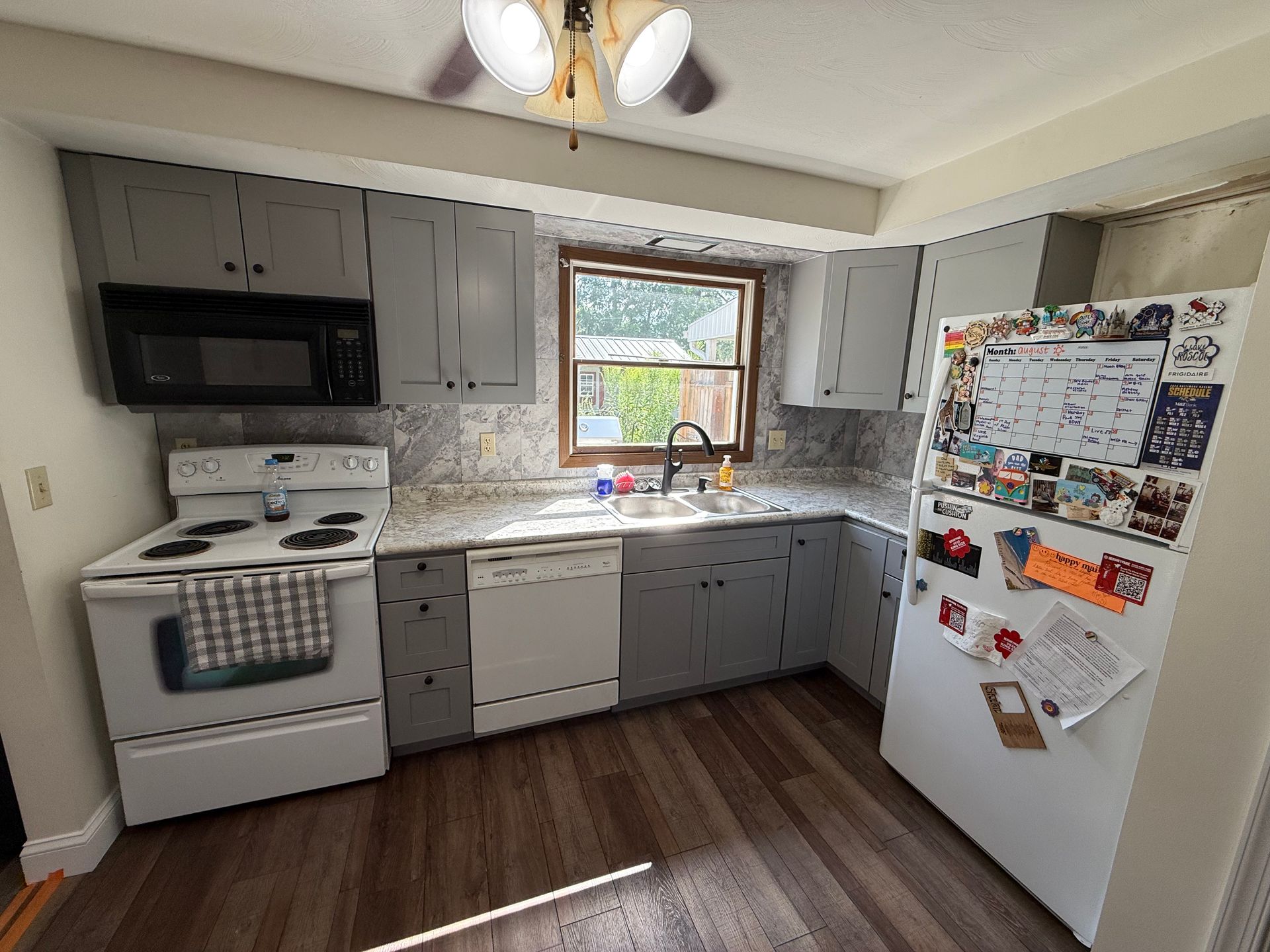 Kitchen with gray cabinets, white appliances, window, and wooden floor. Refrigerator covered with notes.