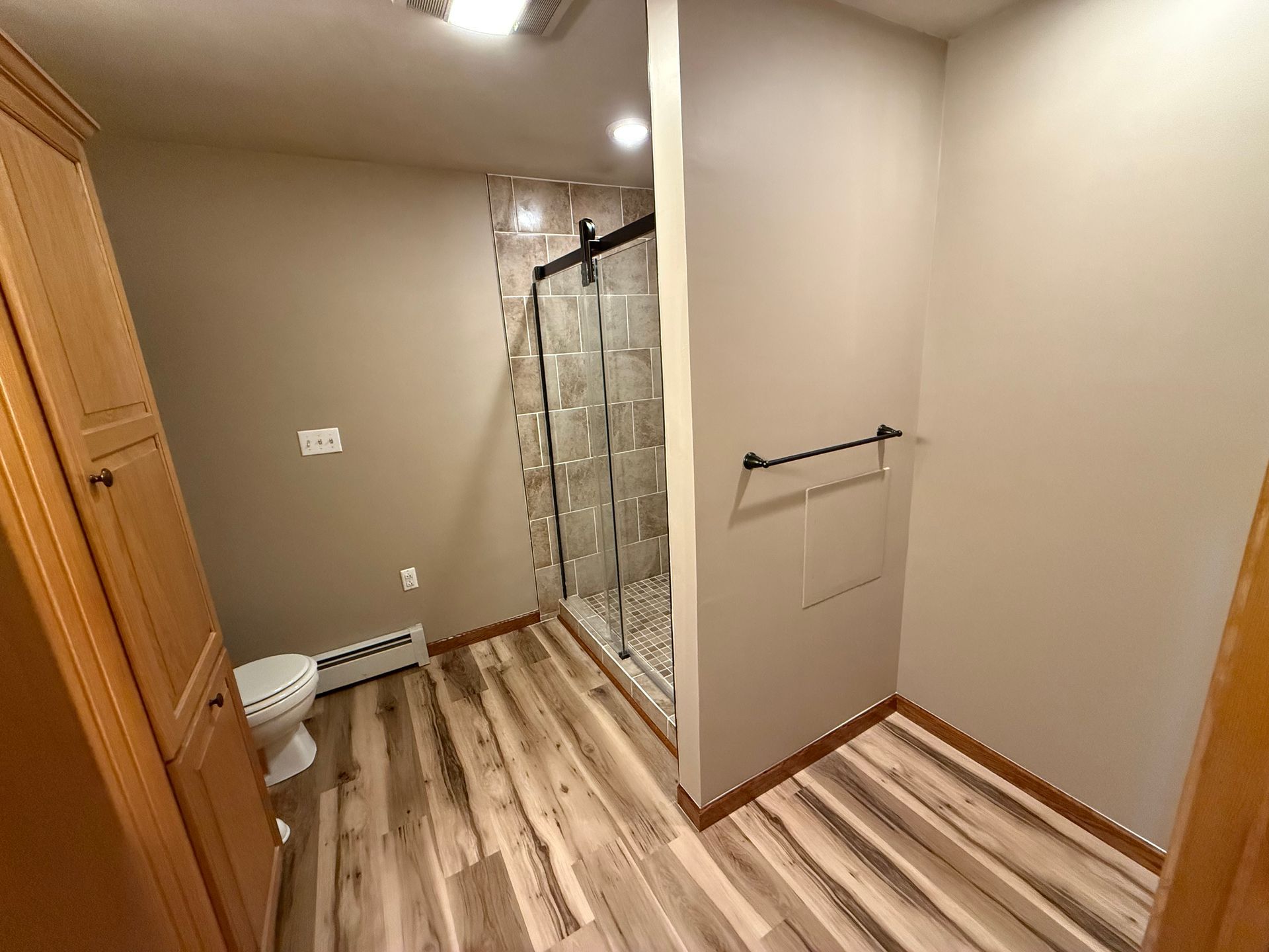 Bathroom with wood-look floor, glass shower, and light brown walls. Tall wooden cabinet and towel rack are visible.