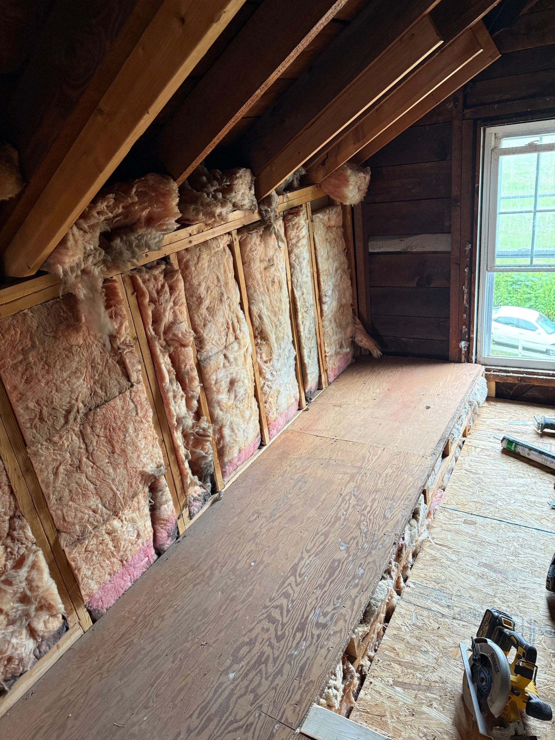 Attic interior with exposed insulation, wooden beams, and a window. A walkway runs along a side wall.
