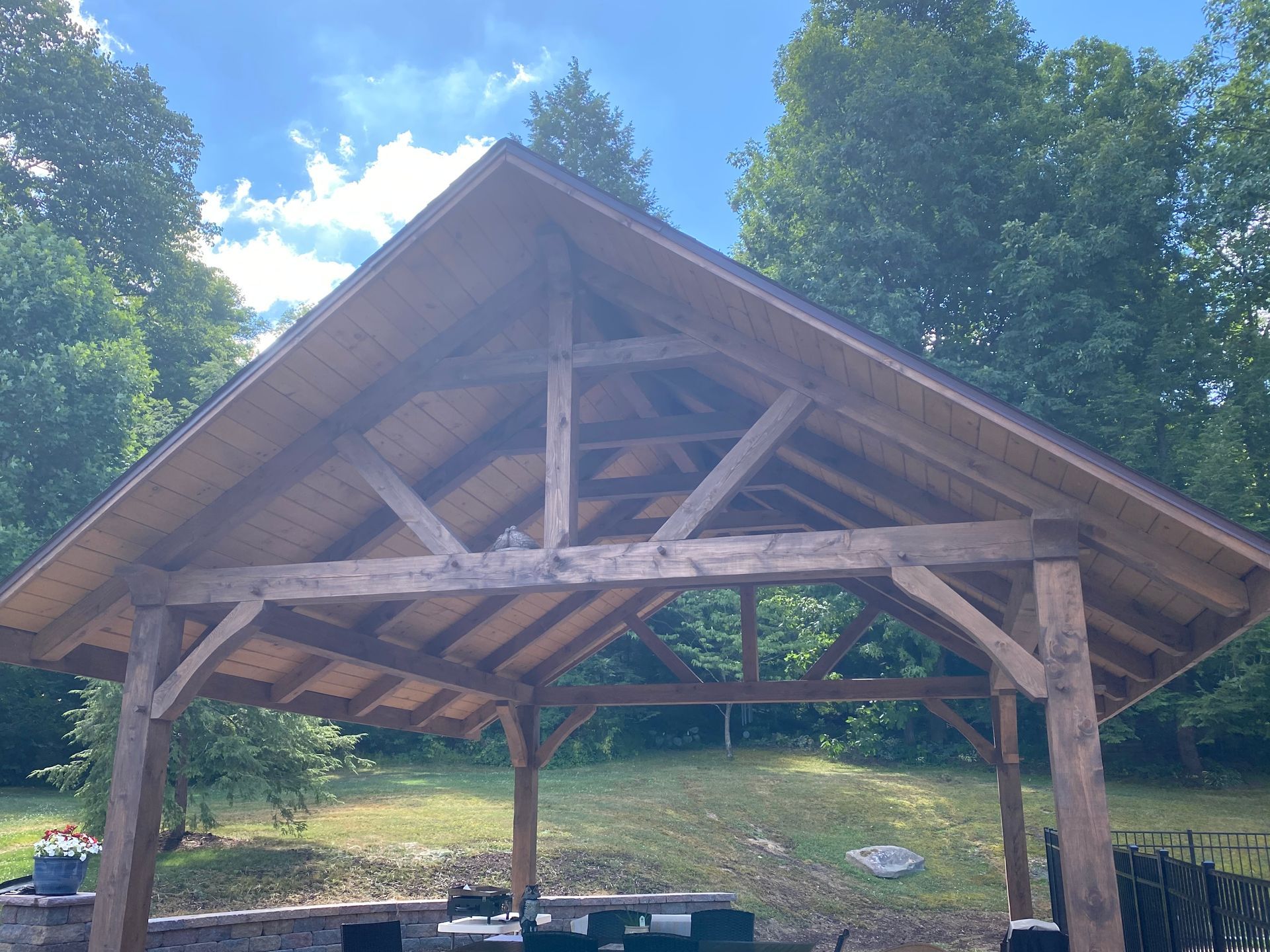 Wooden pavilion with peaked roof, set in a grassy outdoor area under a blue sky.