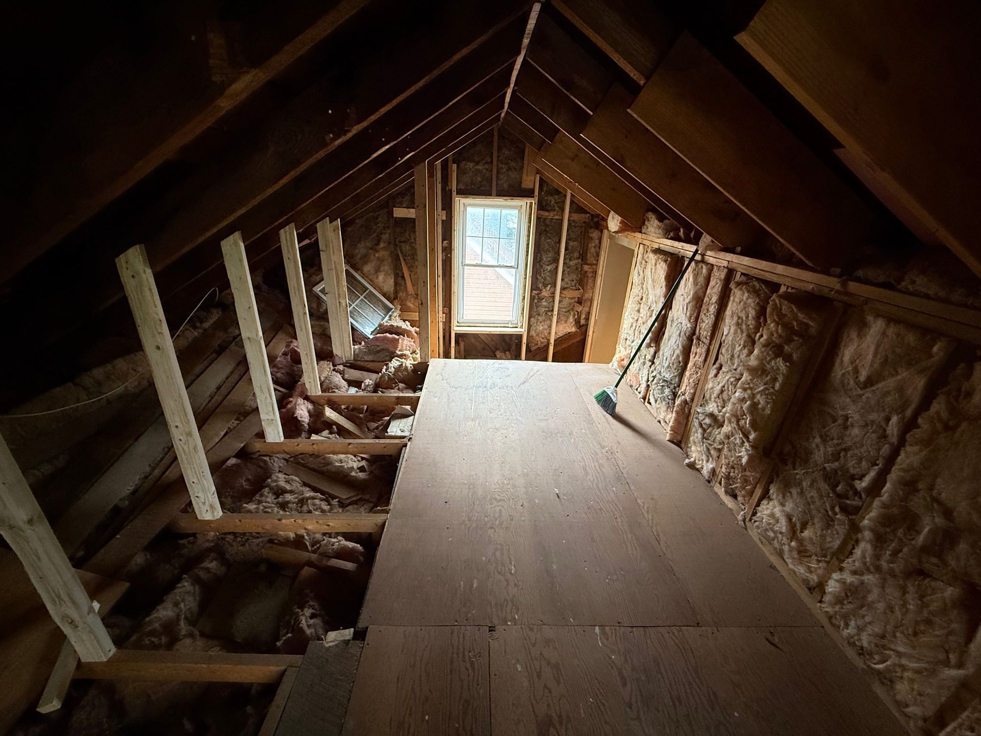 Attic interior with plywood floor, exposed beams, insulation, and a window at the end.