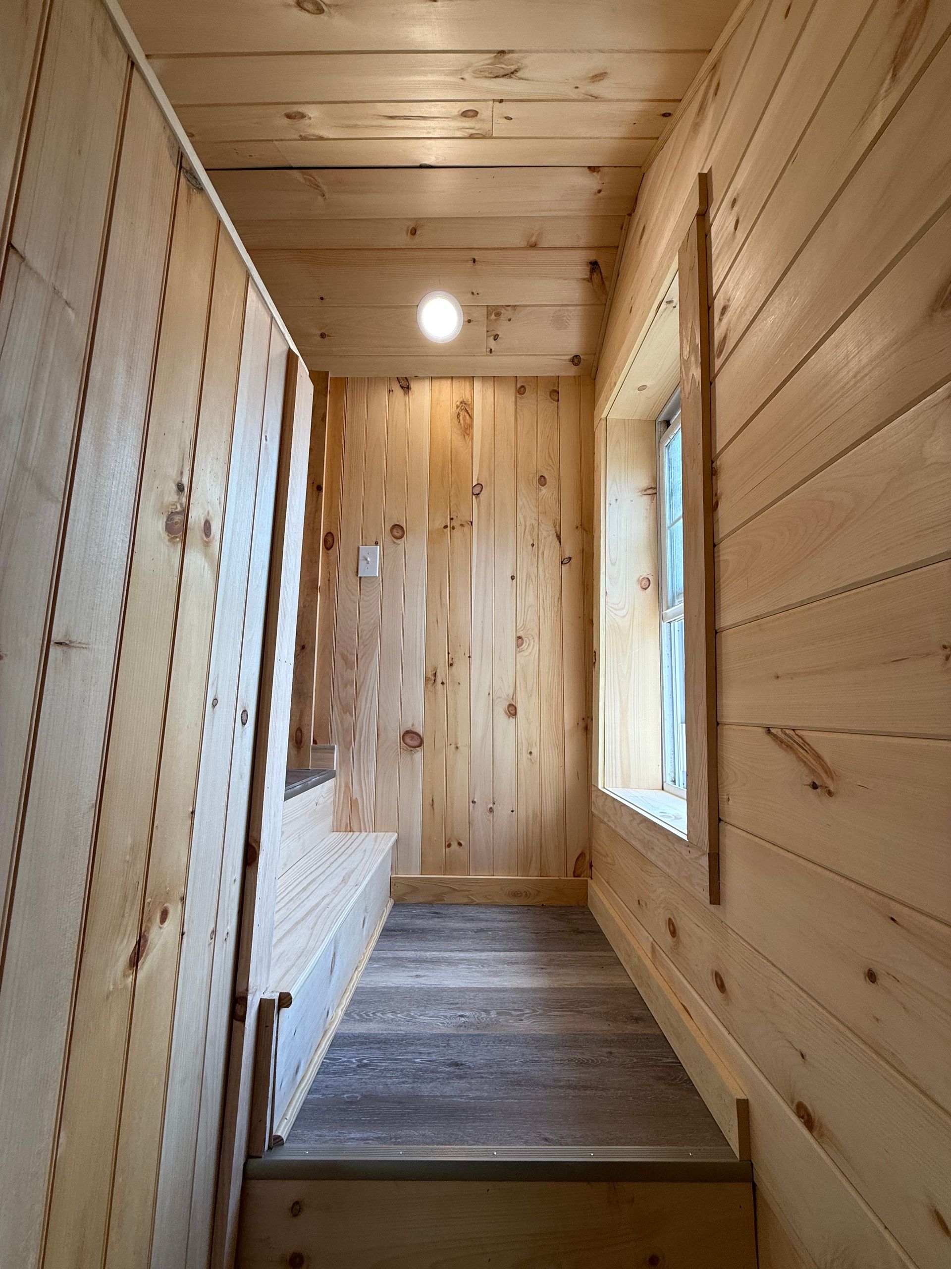 Narrow wooden hallway with steps leading up, wooden walls, small window, overhead light.