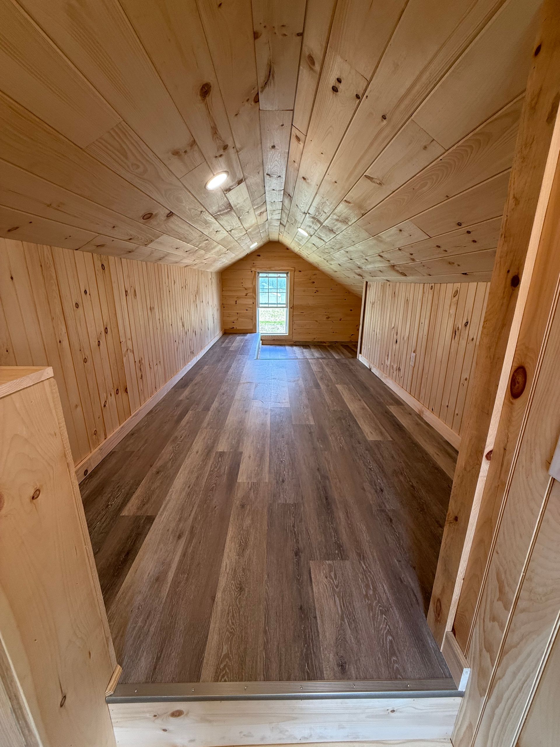 Interior of a small room with wood paneling, flooring, and a window at the end.