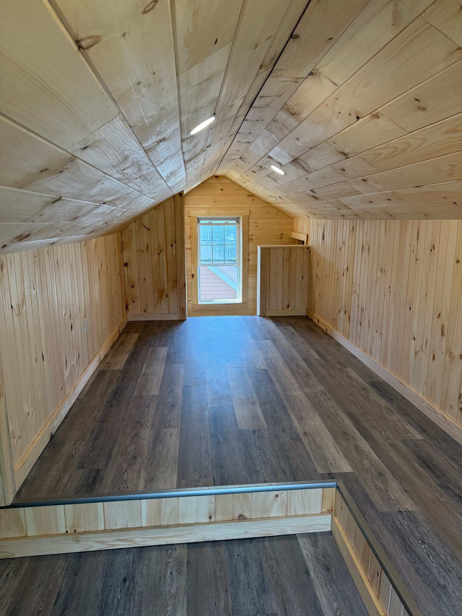 Interior view of a small cabin loft. Wooden walls and ceiling, dark wood floor, small window.
