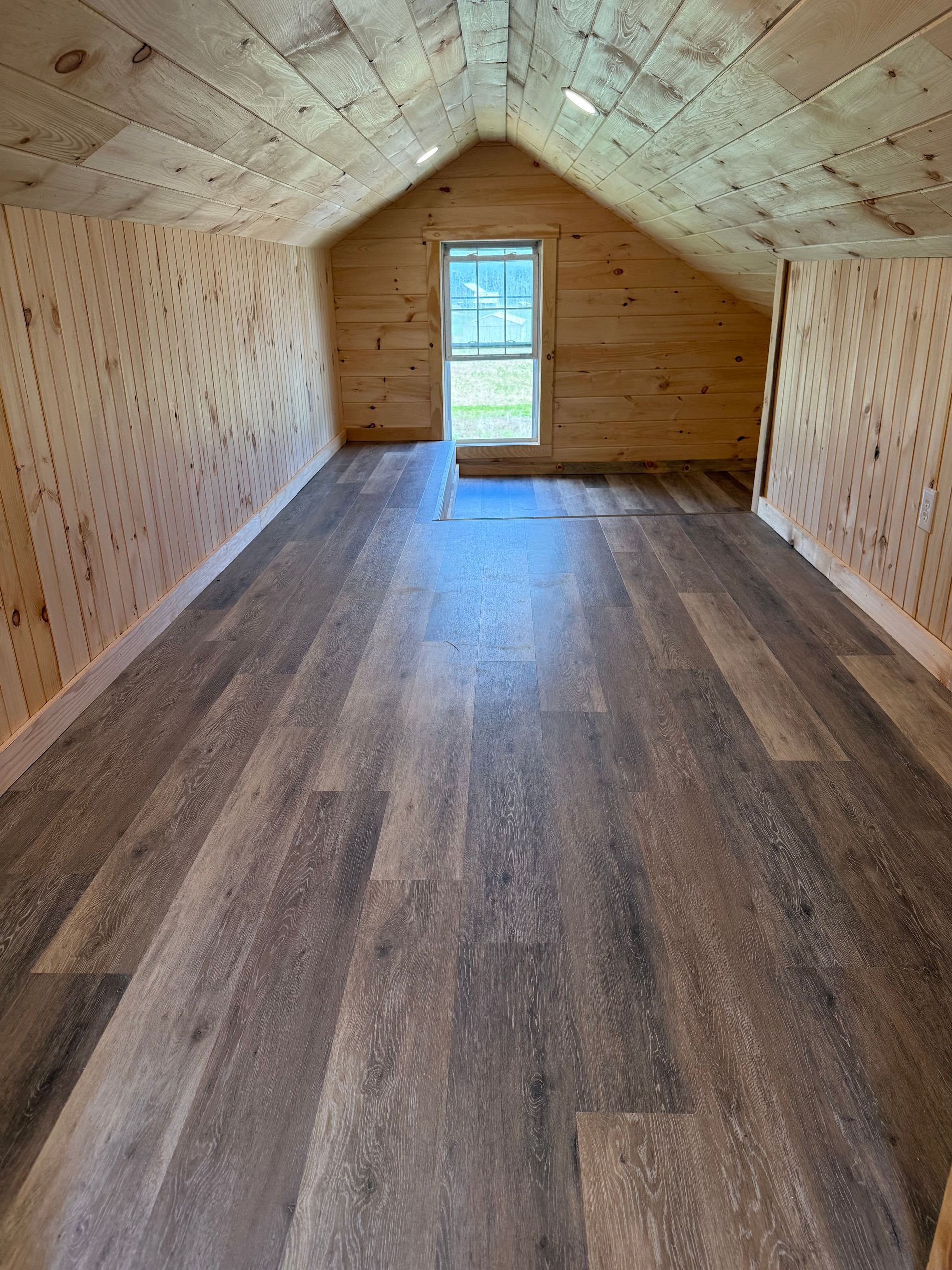 Interior of a small cabin with wood-paneled walls and ceiling, vinyl plank flooring, and a window.