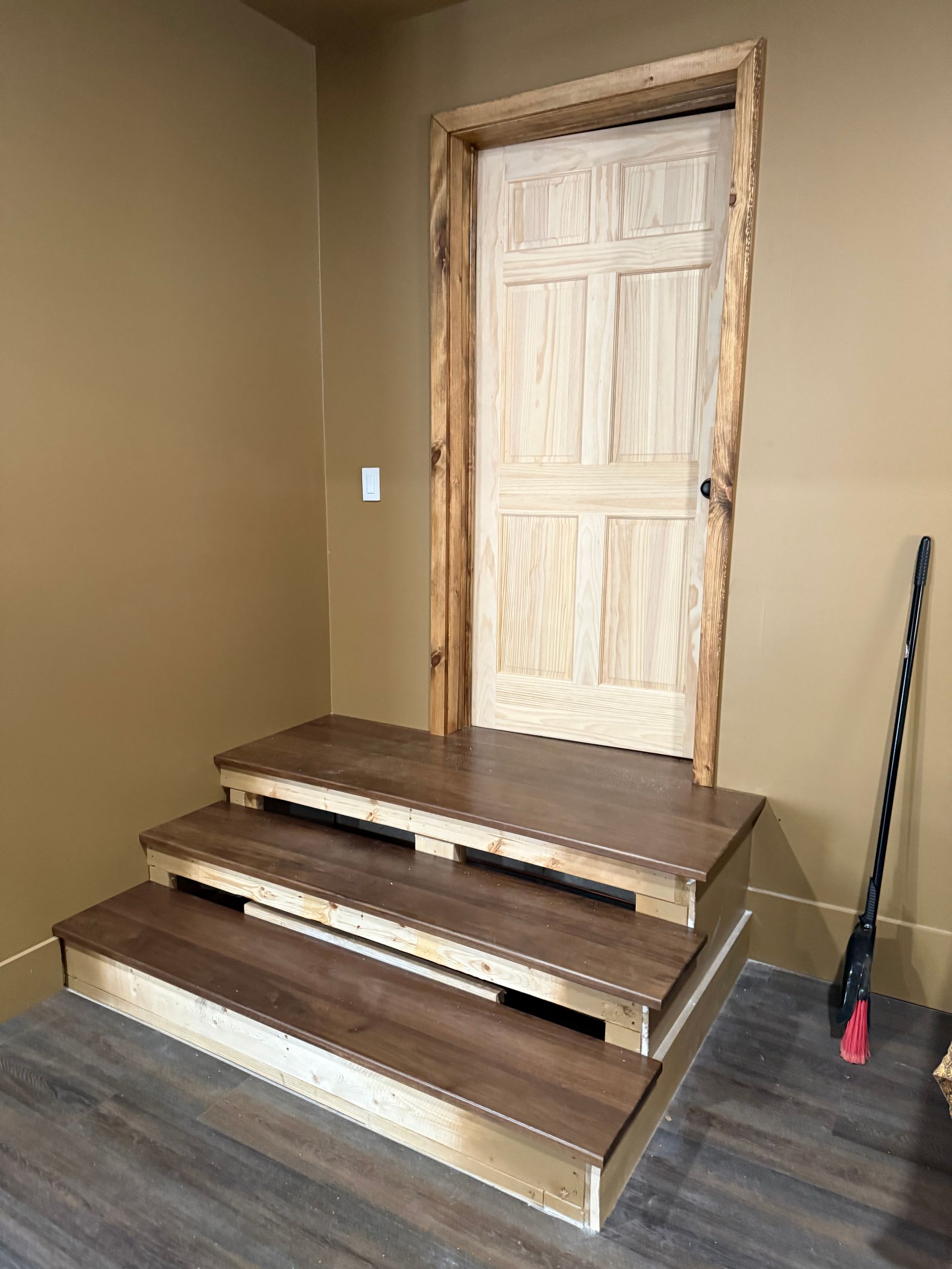 Three brown-topped steps lead to a light-colored wooden door. The doorframe is rustic, and the walls are beige.