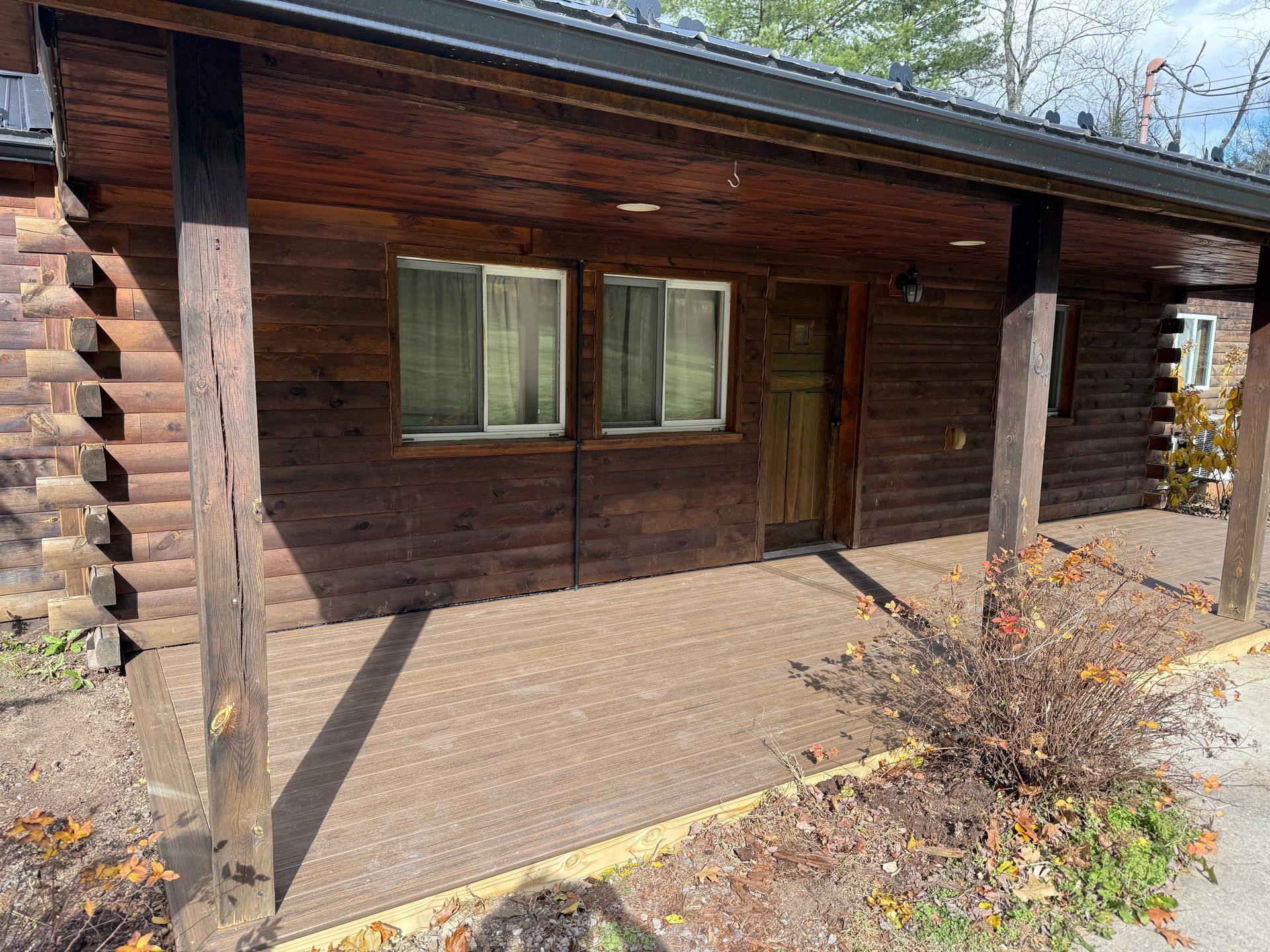 Log cabin with porch, brown wood, two windows, door, and a small bush.