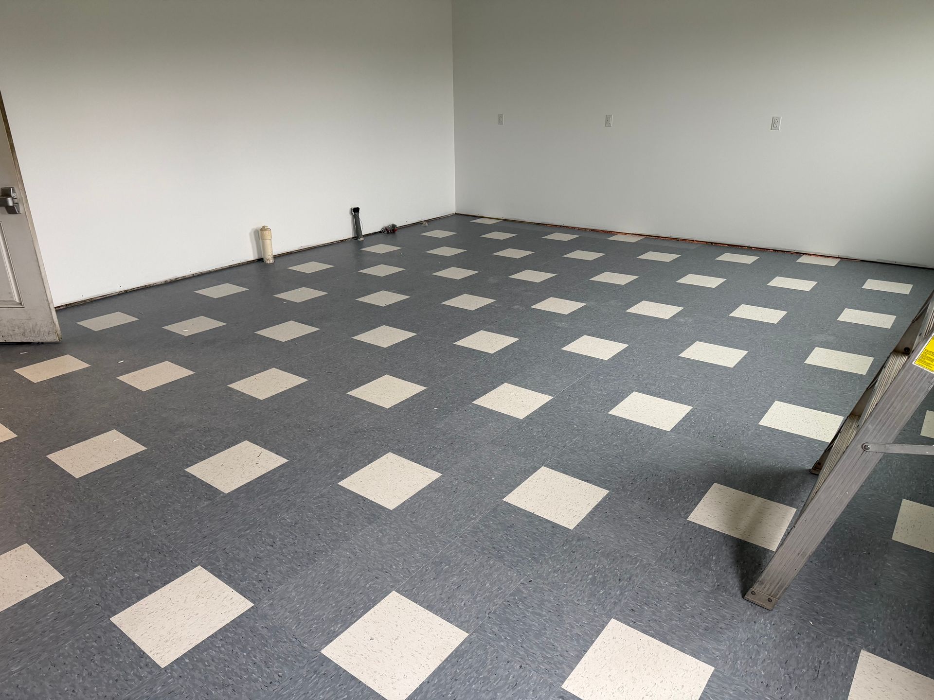 Room with gray and white diamond pattern flooring and white walls; door and pipes visible.