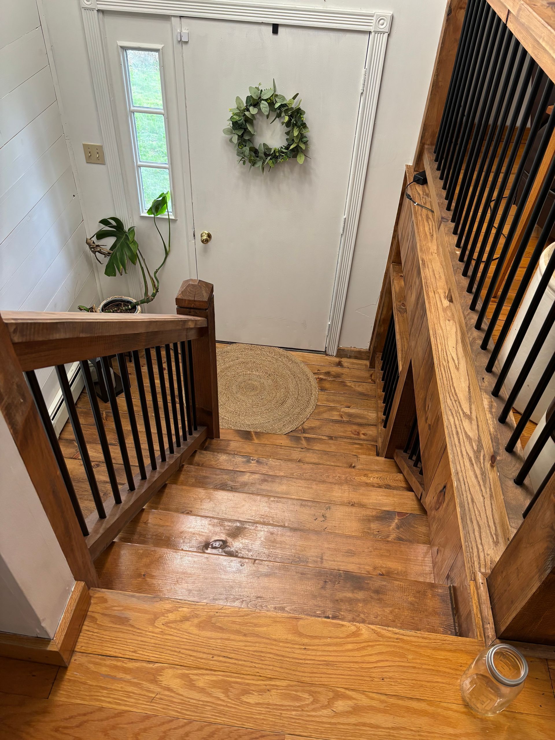 Wooden staircase leading down to a white door with a wreath; a houseplant and a rug are visible.