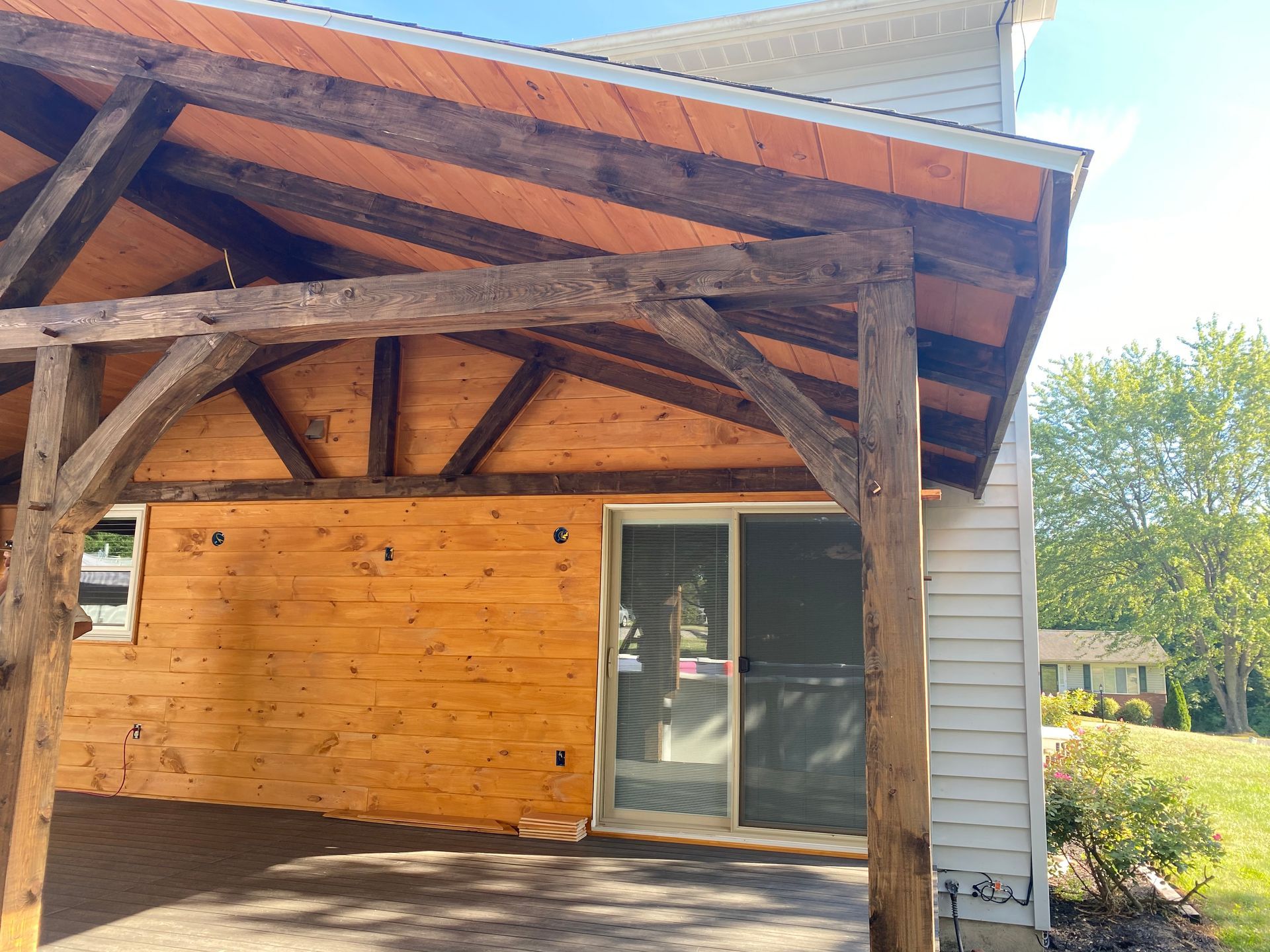 Wooden-framed covered patio attached to a house with a sliding glass door and stained wood ceiling.