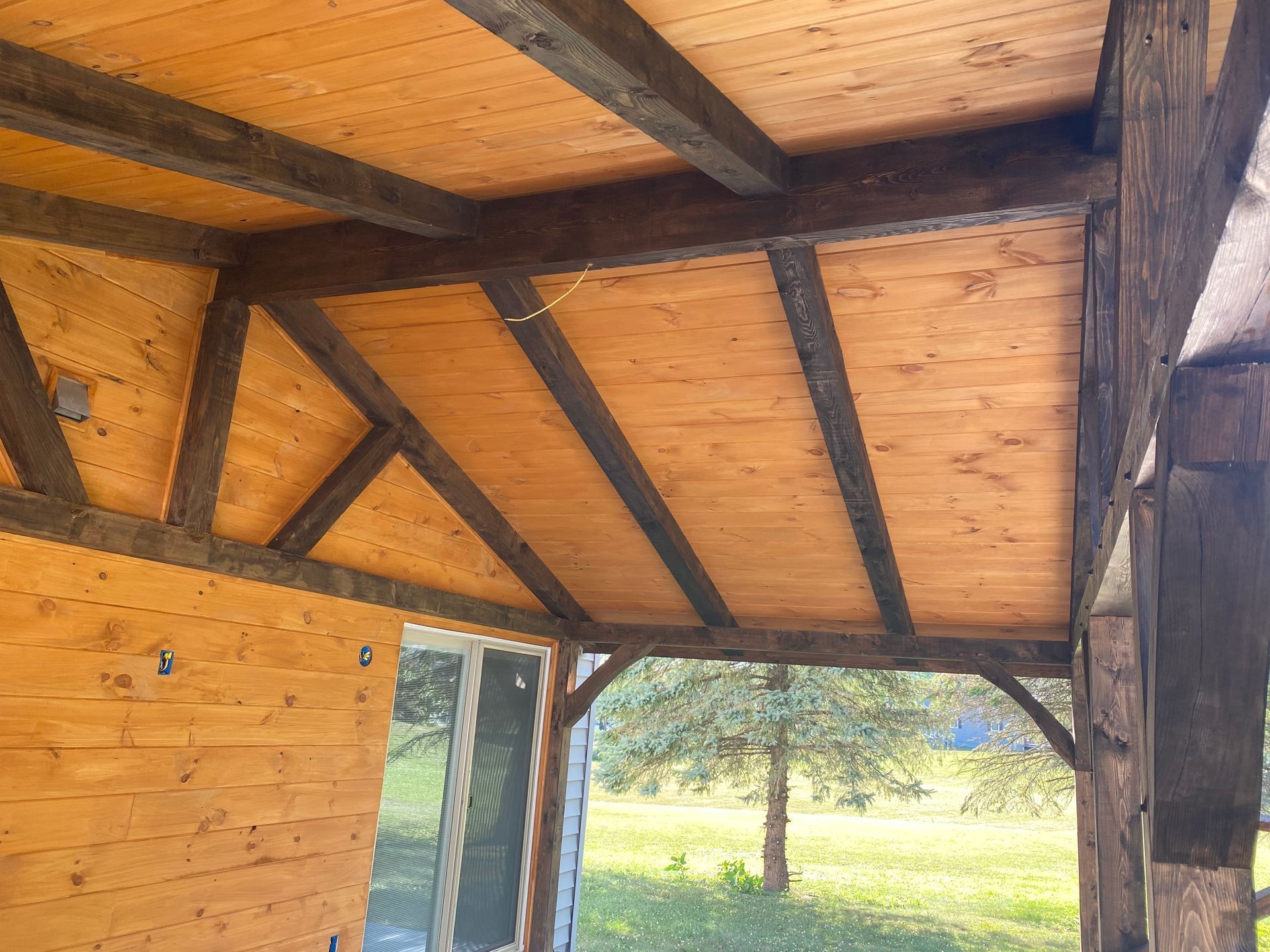Wooden porch roof with dark beams and light wood panels, overlooking a green yard and tree.