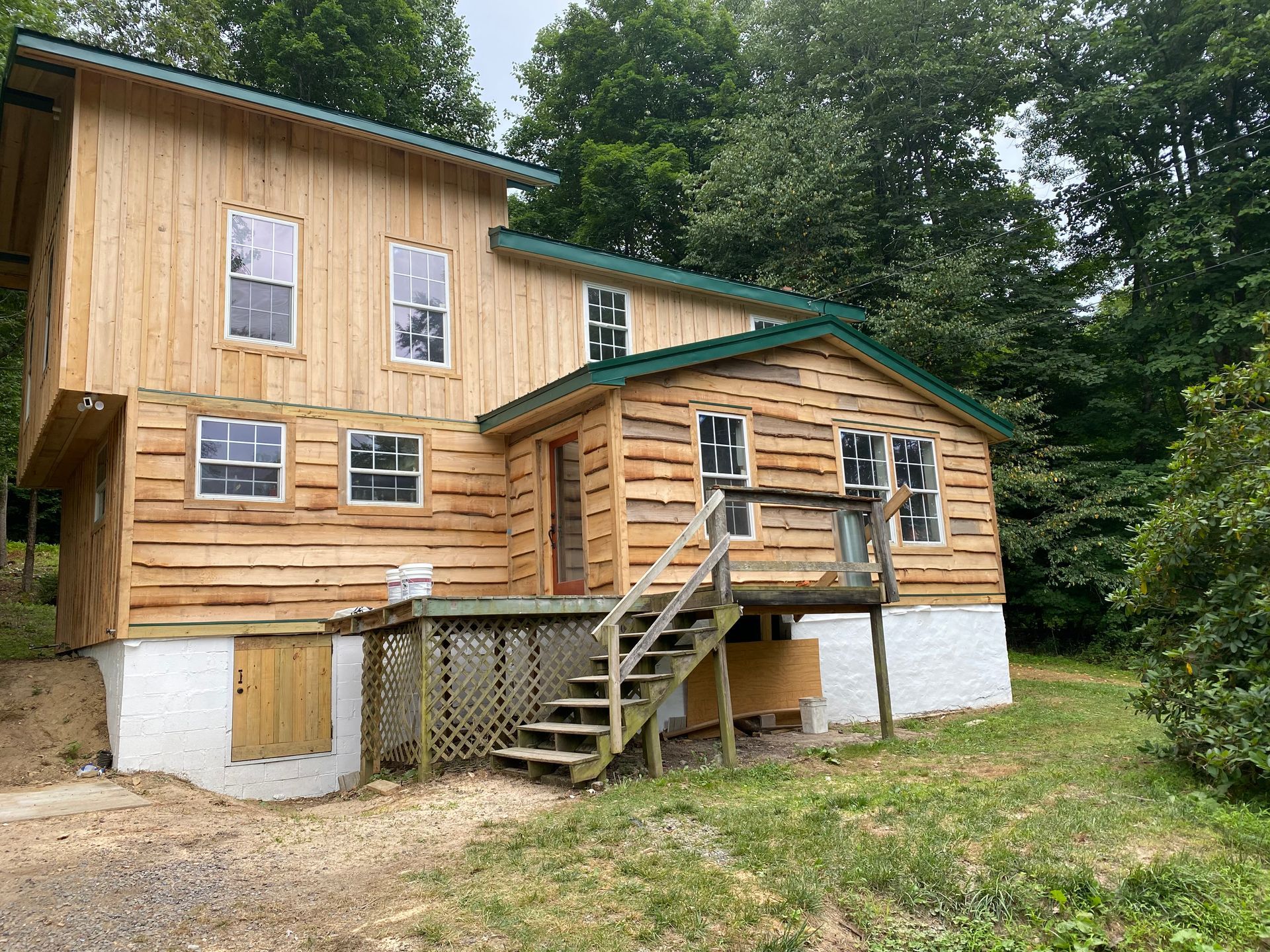 Two-story wooden house with a small porch and stairs. Surrounded by trees and grass, on a slight hill.