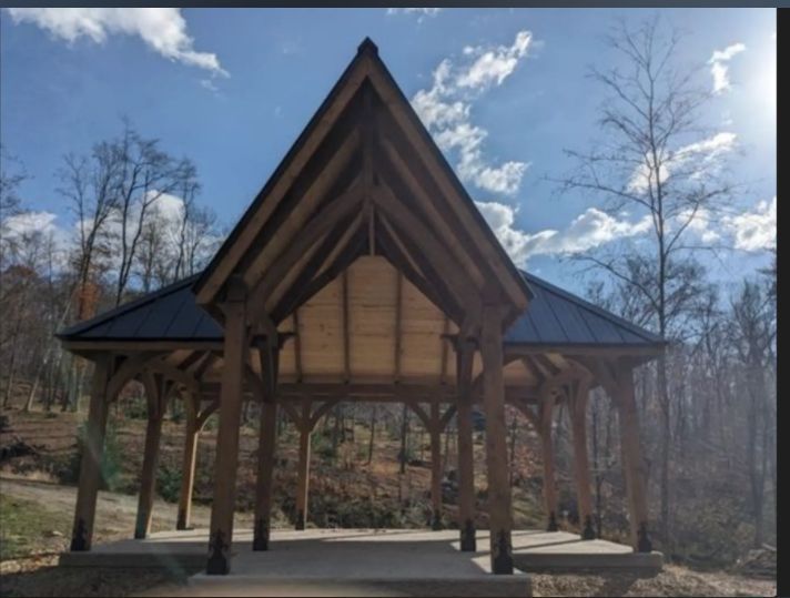 Wooden gazebo with a dark roof in a wooded area on a sunny day.