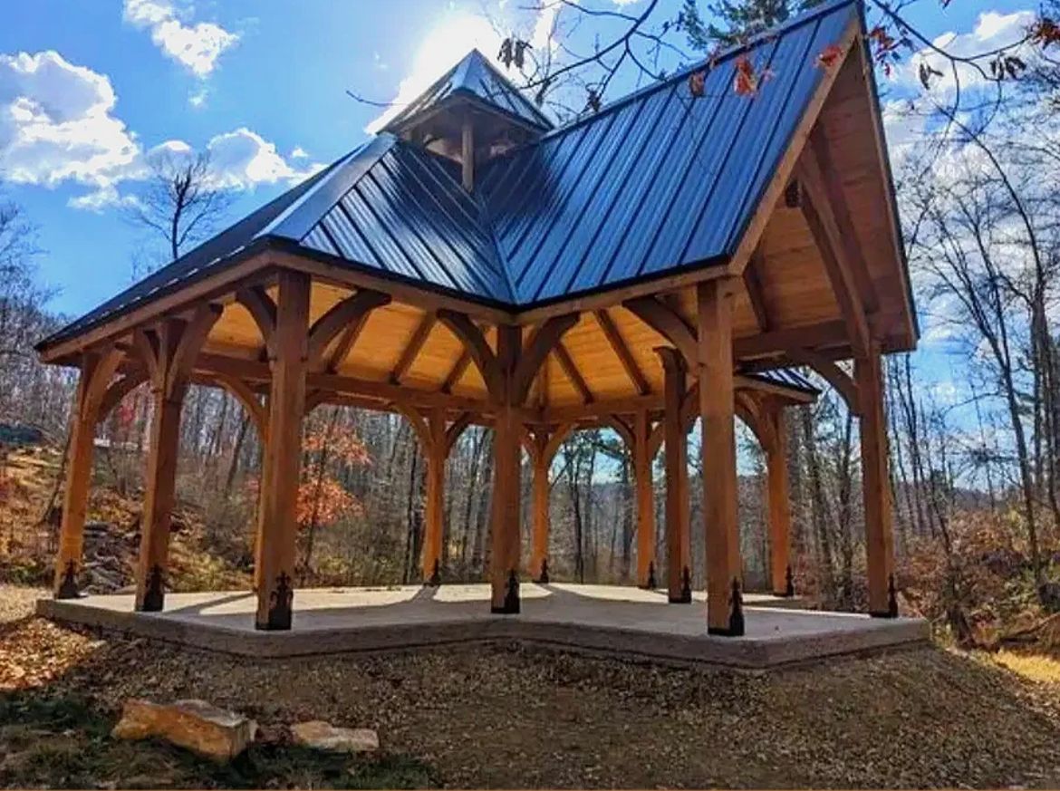 Wooden gazebo with black roof on a concrete pad, surrounded by trees and a sunny sky.