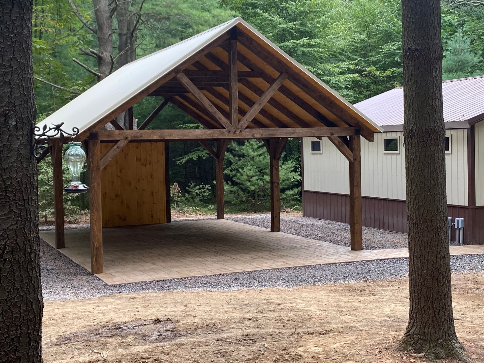 Wooden carport with metal roof, set on gravel, next to a building and trees.