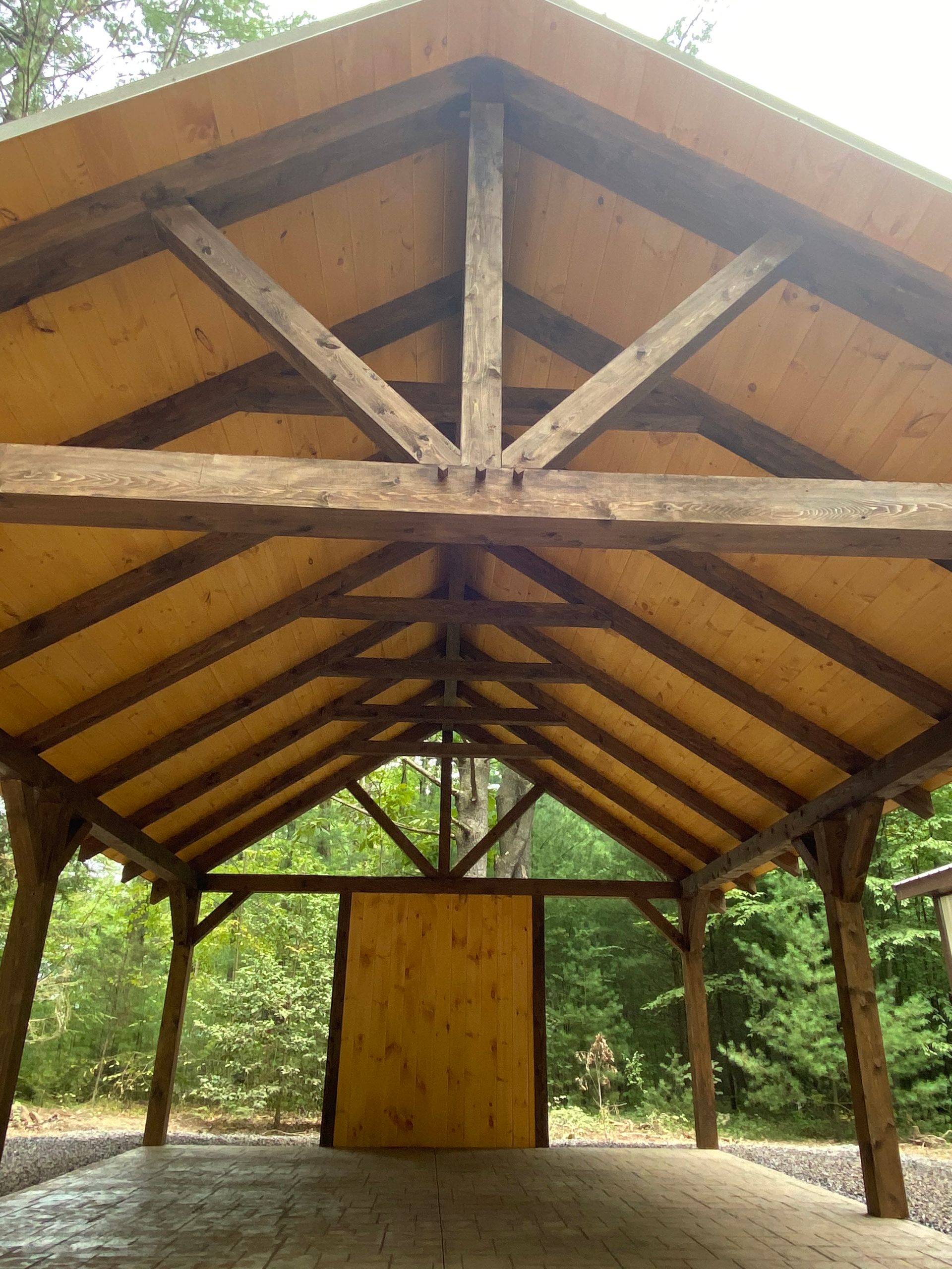 Wooden roofed pavilion with support beams and a wooden door, set in a forest.