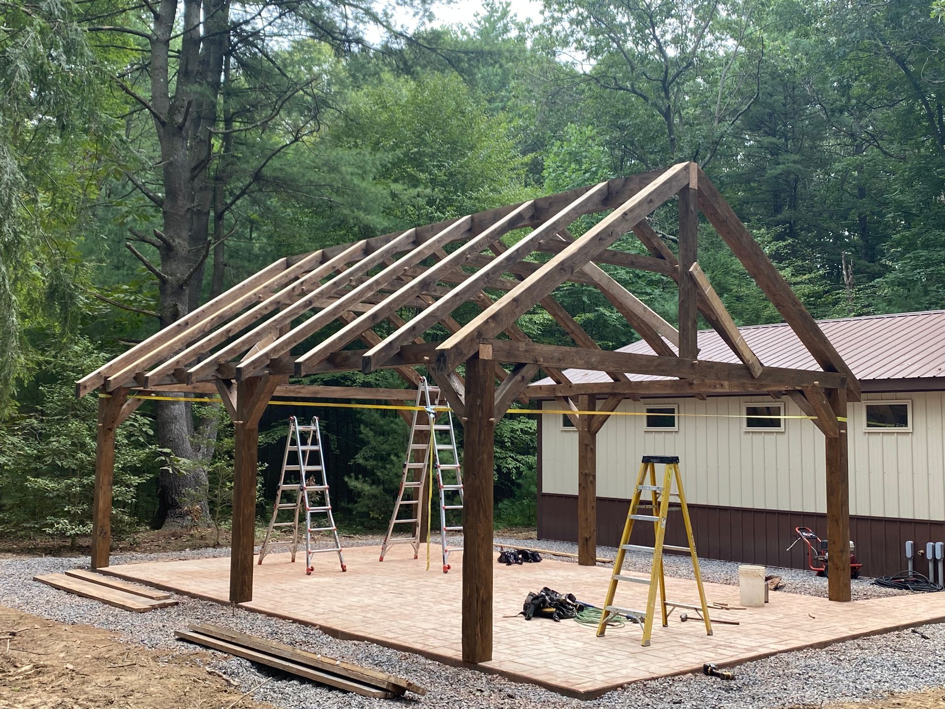 Wooden pavilion under construction, framing a gravel area with ladders and building materials.