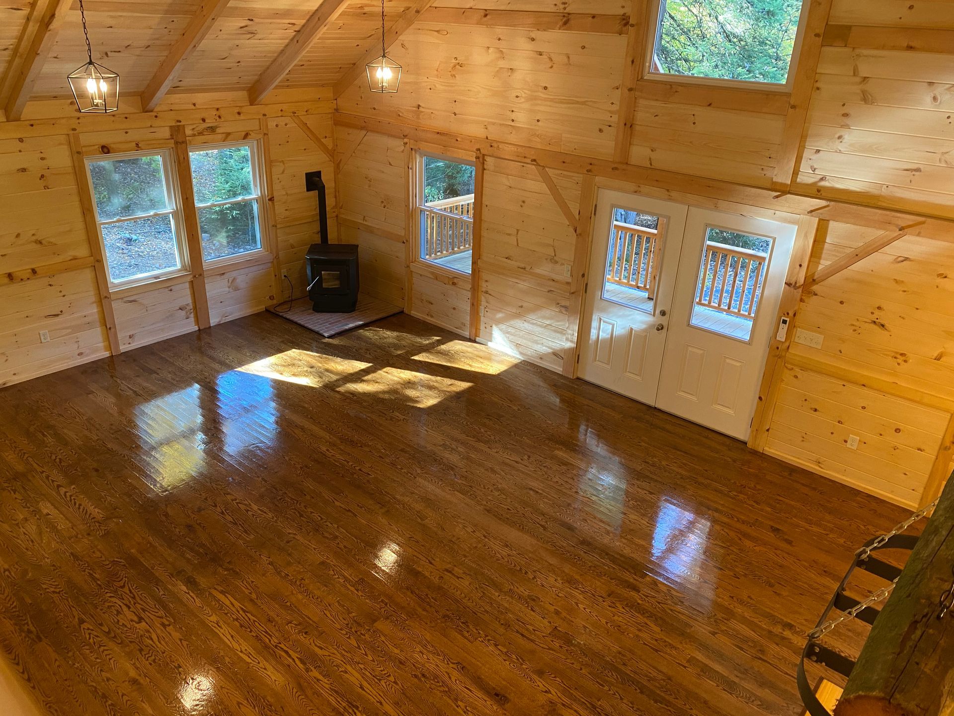 Interior of a wooden cabin with shiny hardwood floors, windows, and a wood-burning stove.