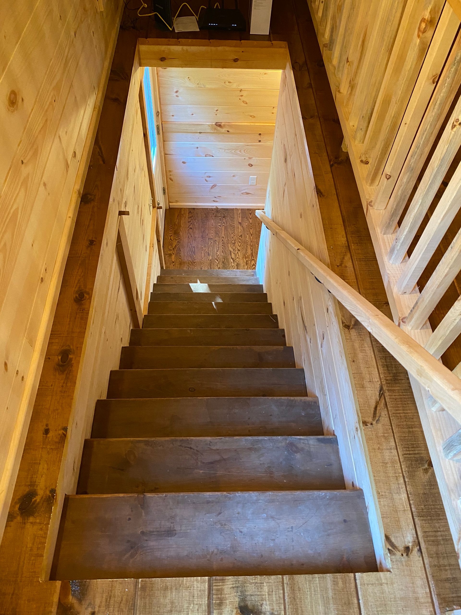 Wooden staircase leading down into a rustic, wood-paneled room. Handrail on right side.