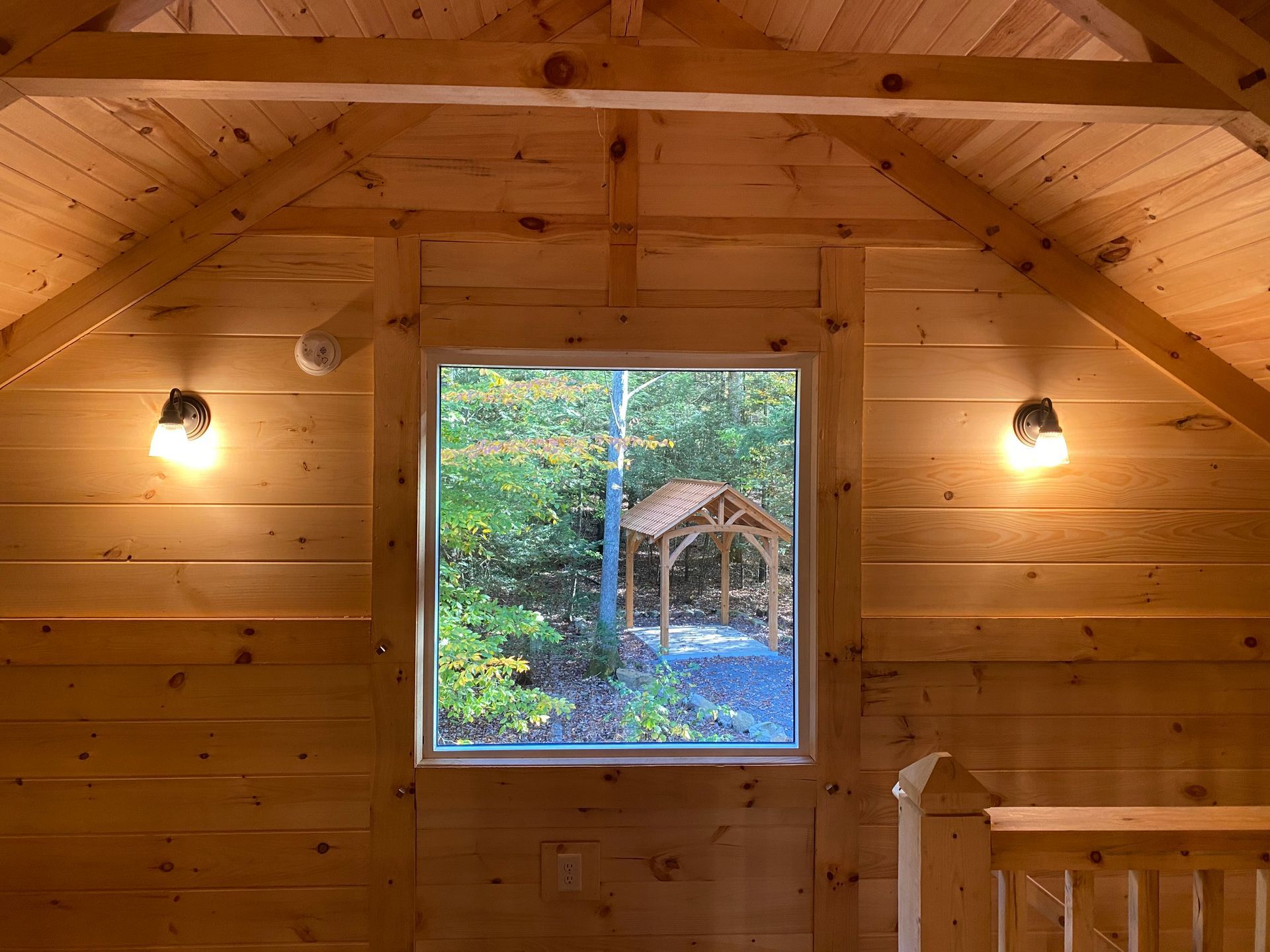 Interior of wooden cabin with window overlooking a forest and a small wooden structure; two wall sconces.