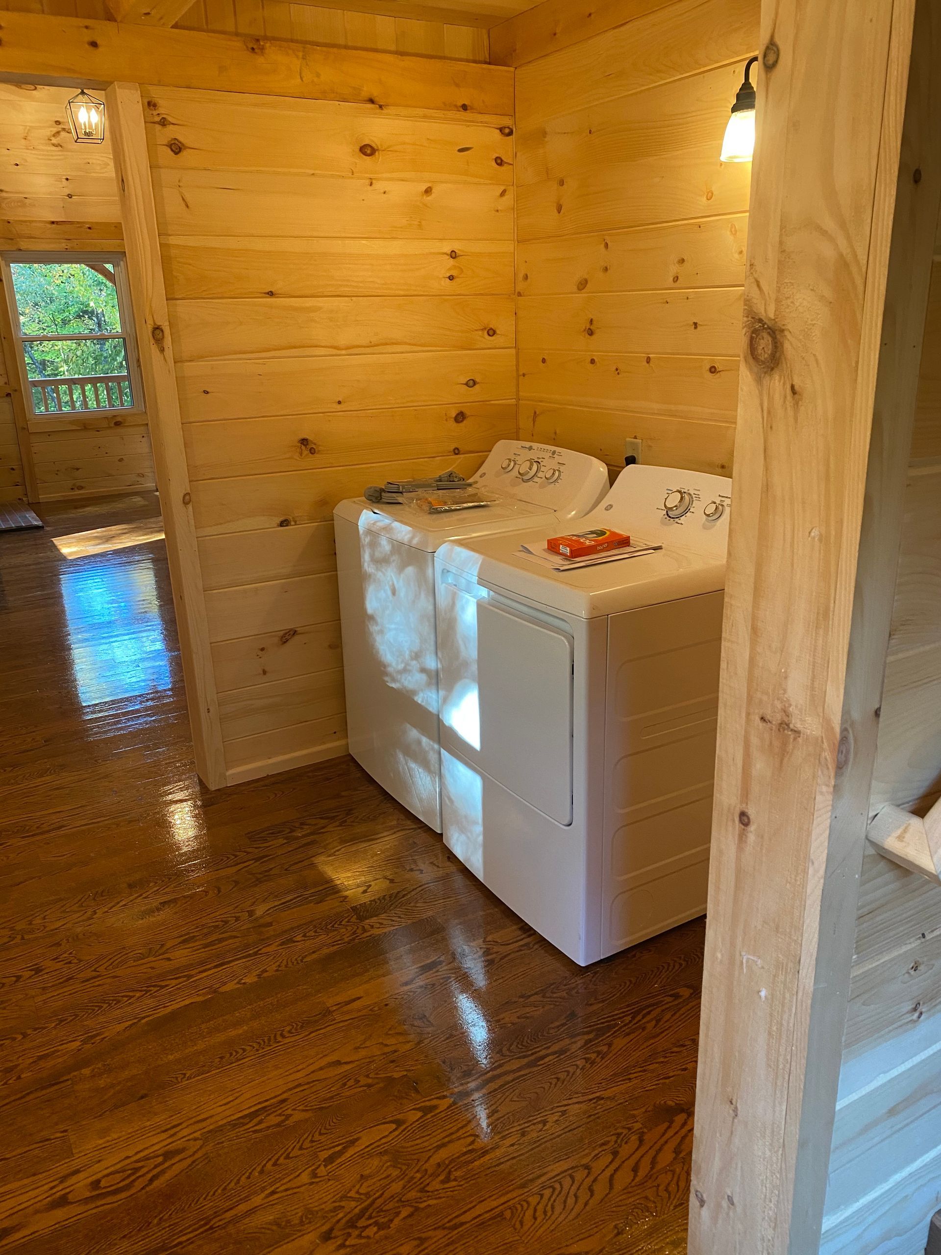 Washer and dryer in a wood-paneled room.  Shiny wooden floor, natural light from a window.