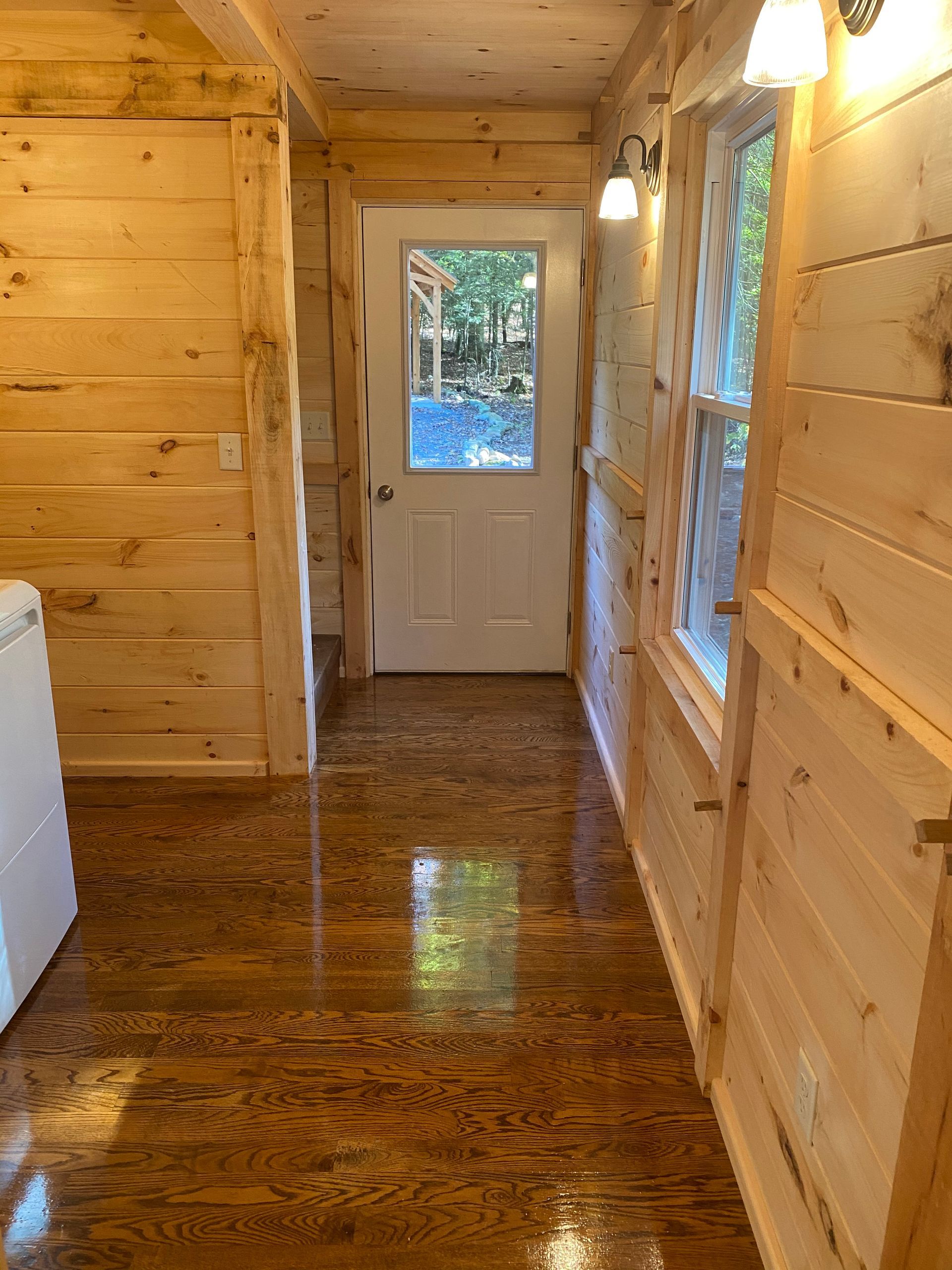 Wooden-walled hallway with door at the end and a window to the right. The floor is shiny and reflective.