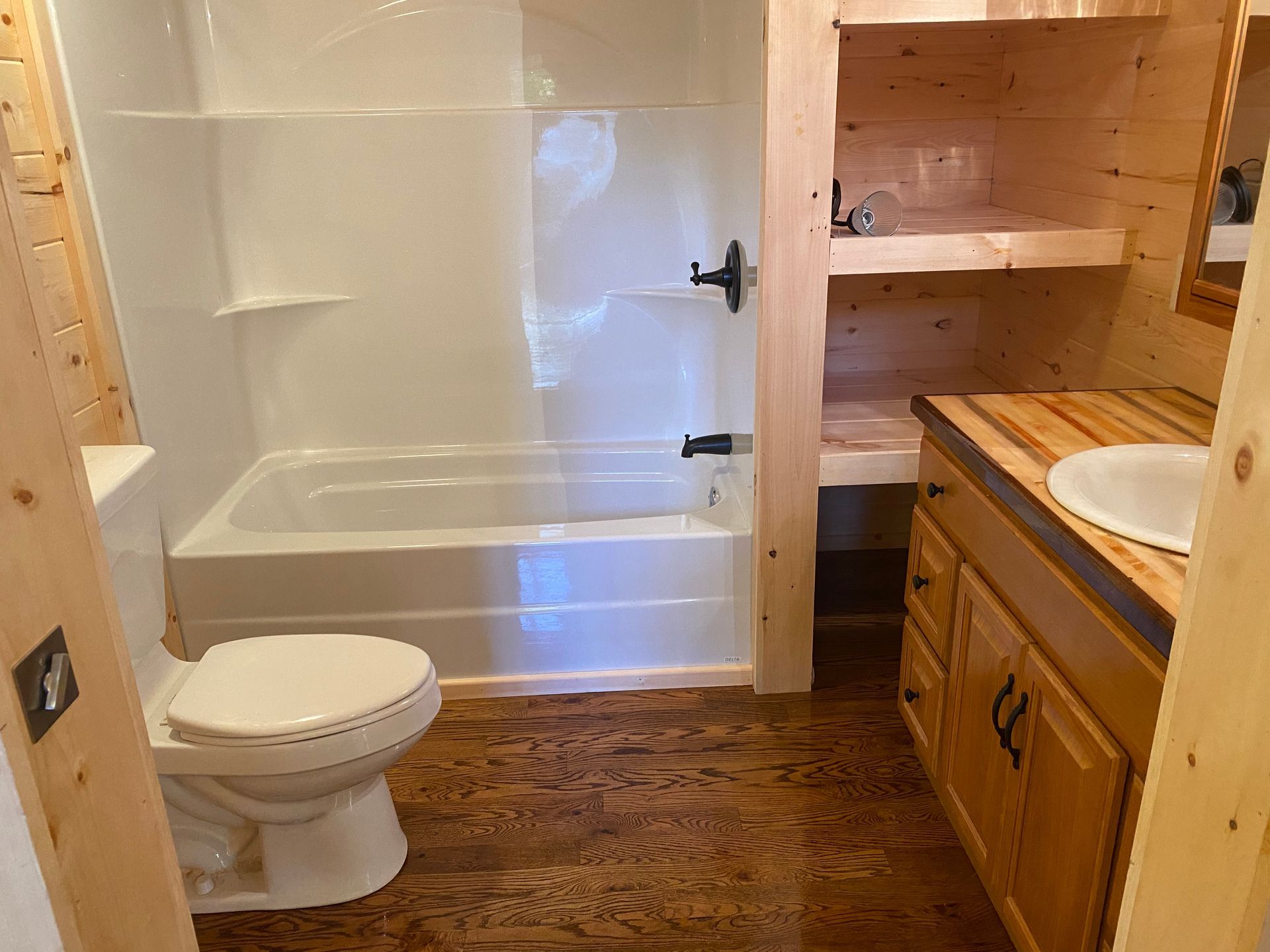 Bathroom with white tub/shower, toilet, wood vanity, and shelves; wood floors and walls.