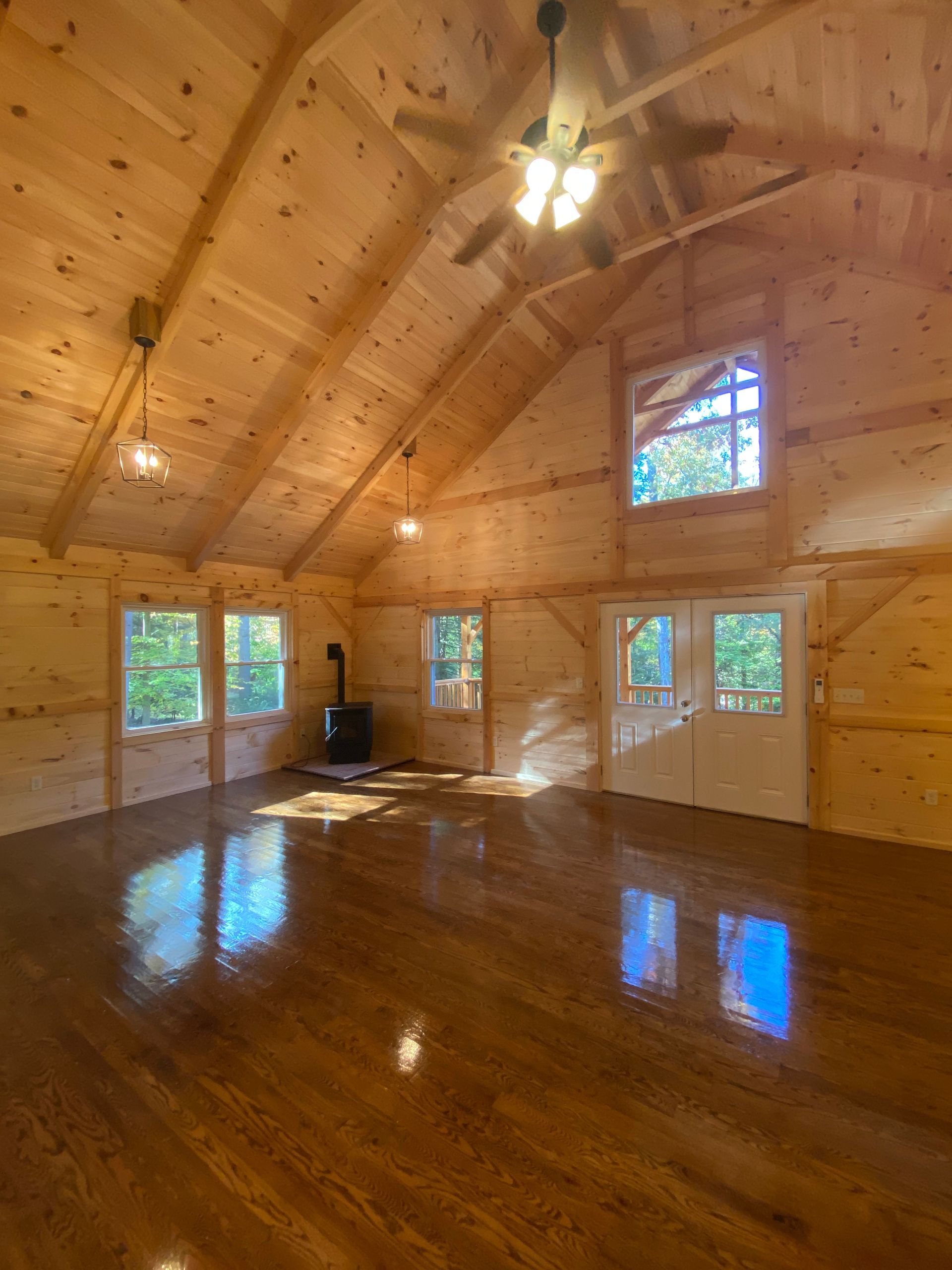 Interior of a wooden cabin with a wood stove, windows, glossy wood floor and ceiling, and a ceiling fan.