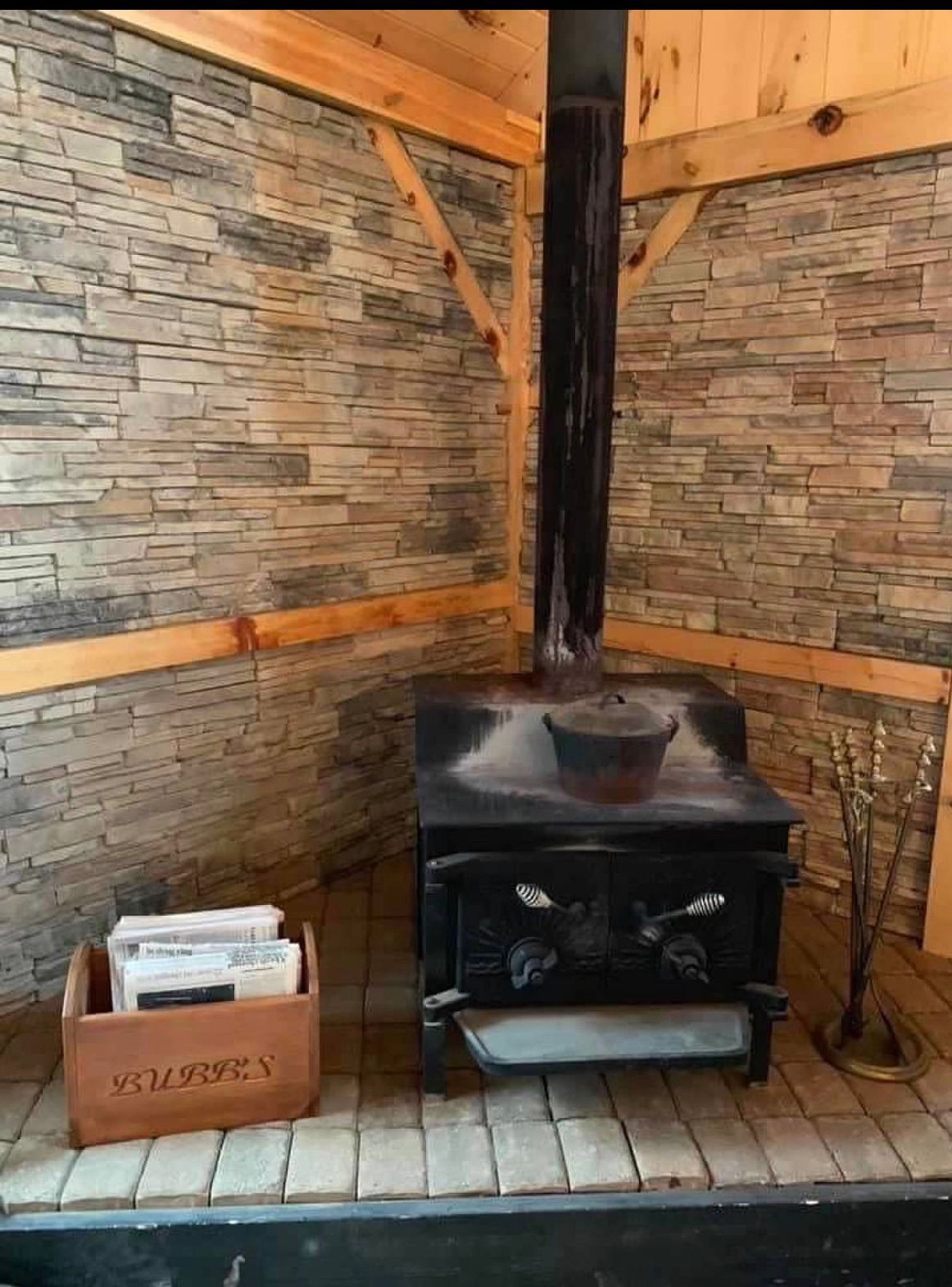 Wood-burning stove in a rustic room with stone wall and wooden beams. A wooden box and metal chimney are present.