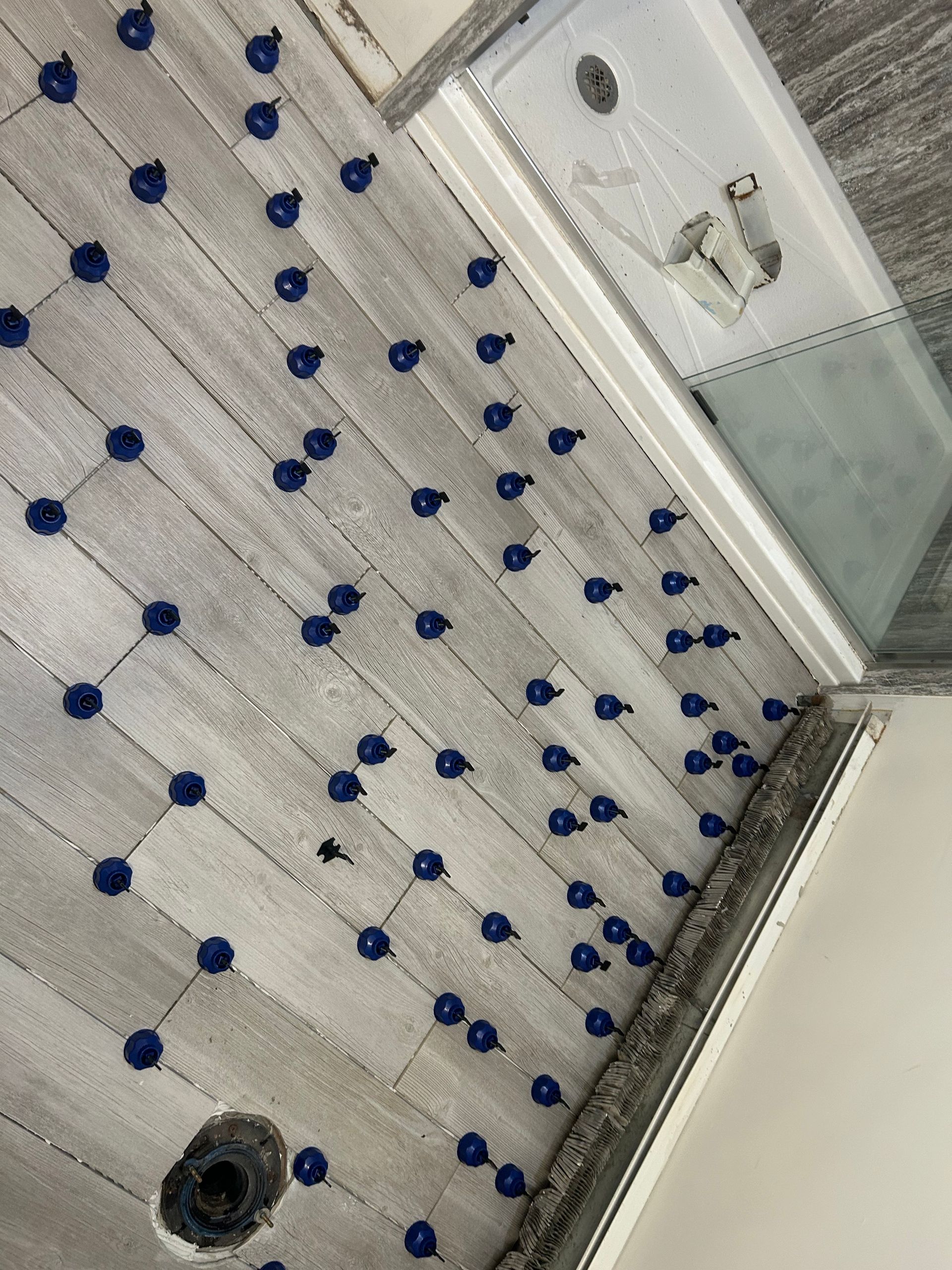 Bathroom floor with light grey wood-look tiles, blue leveling spacers, and a partially built shower.