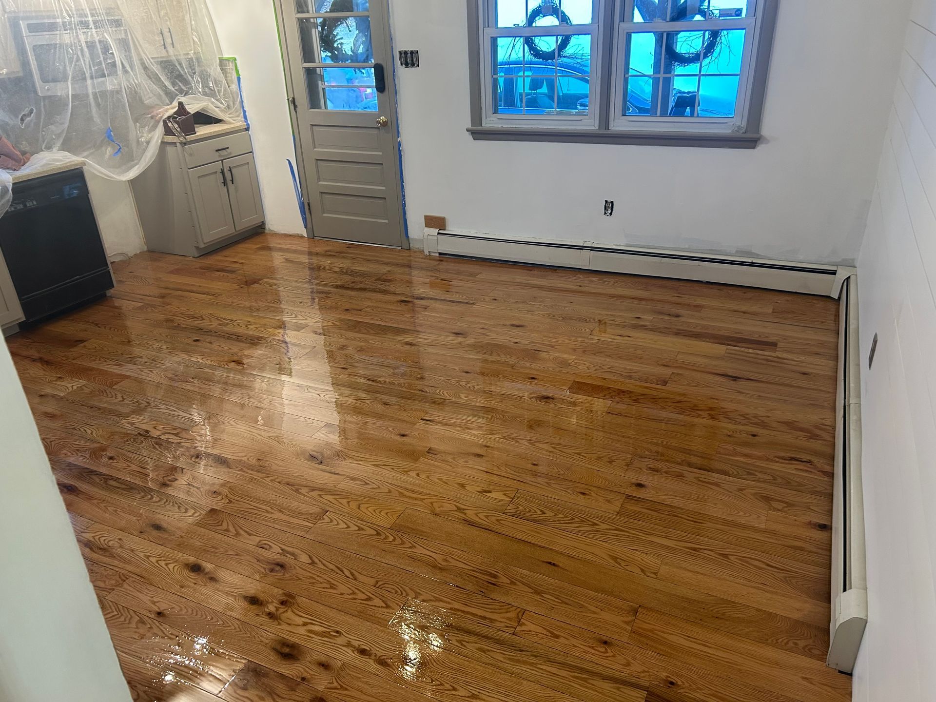 Hardwood floor in a room with a window, door, and cabinets; partially covered in plastic.