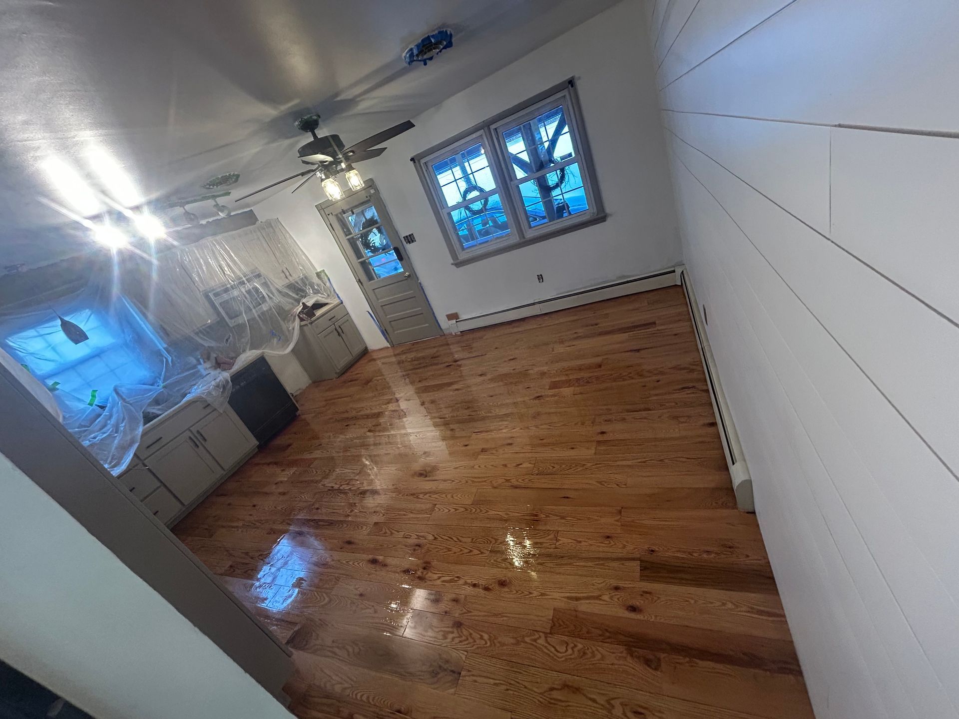 Hardwood floors in a room with white paneled walls, a kitchen with covered appliances, and light fixtures.