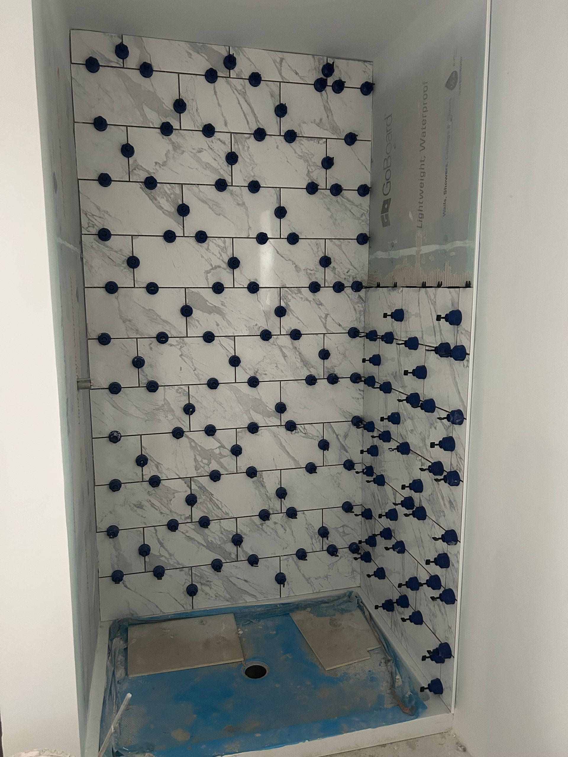 Shower stall with white marble-like tiles being installed; blue leveling system used to space tiles; light and shadow.