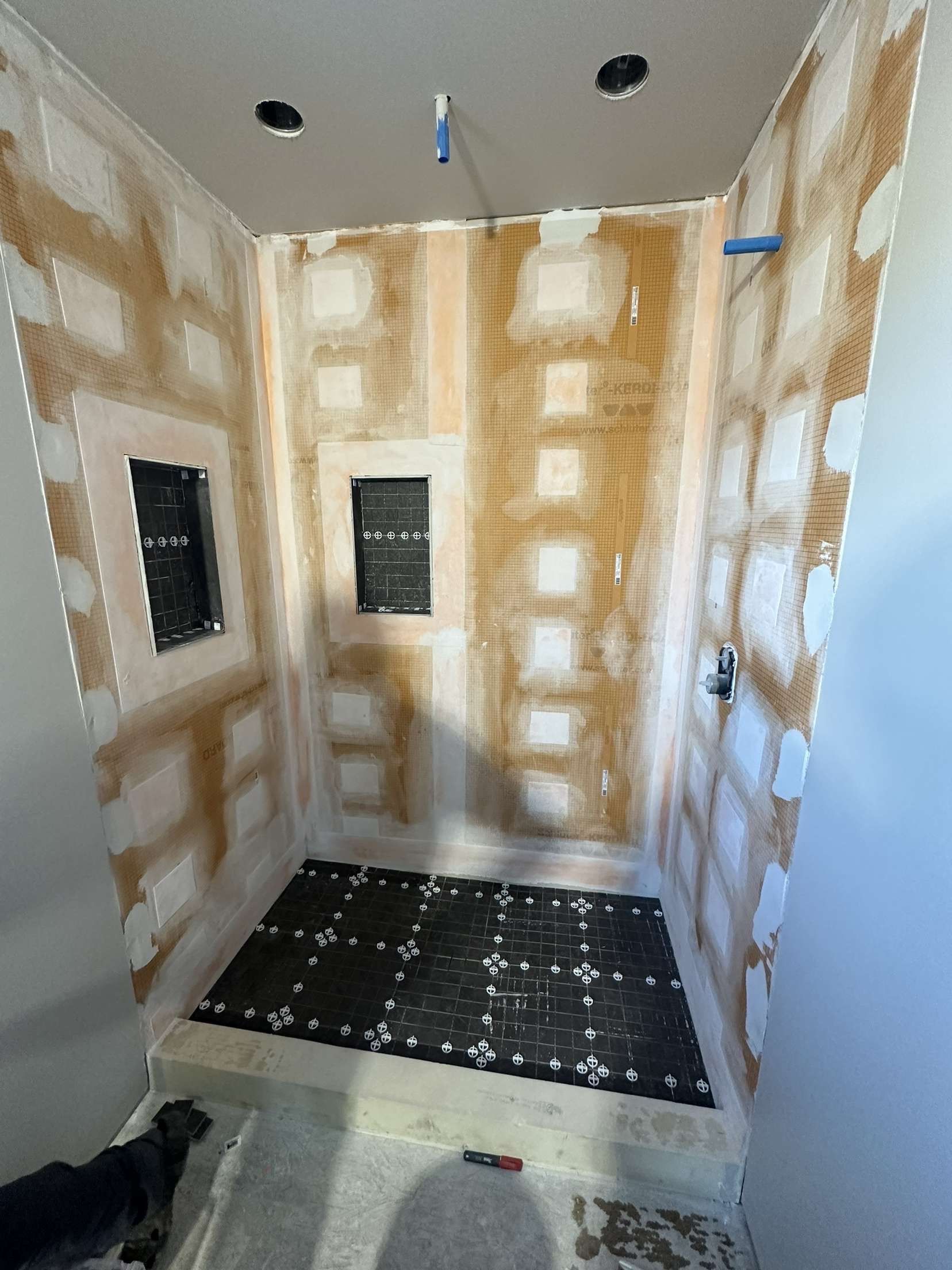 Shower stall under construction, beige and white walls, black floor tile, dog on the edge.