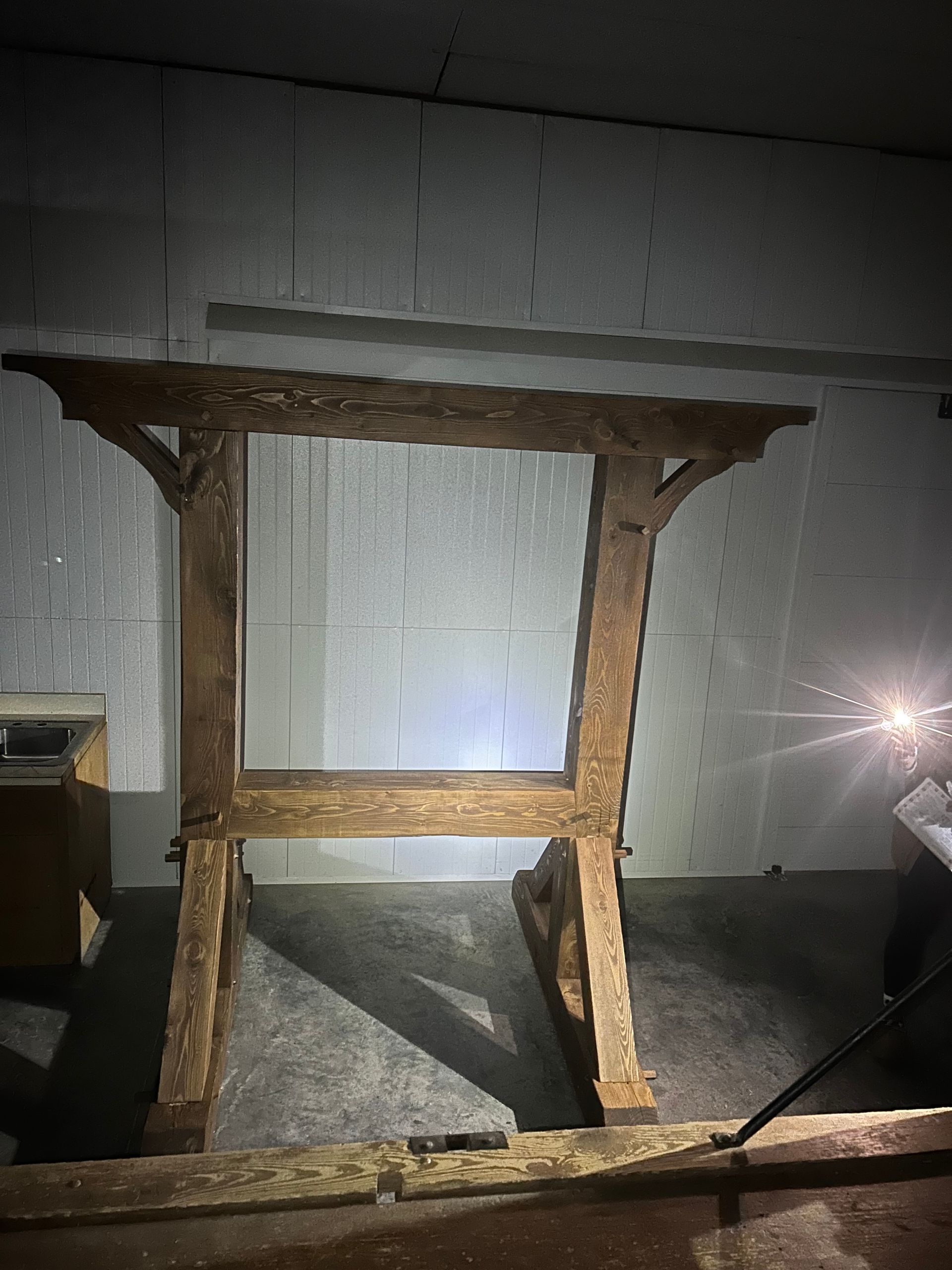 Wooden frame structure, possibly for a door or window, in a dimly lit indoor space, casting a shadow.