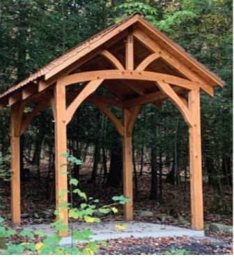 Wooden gazebo in a forest, featuring arched supports and a peaked roof.