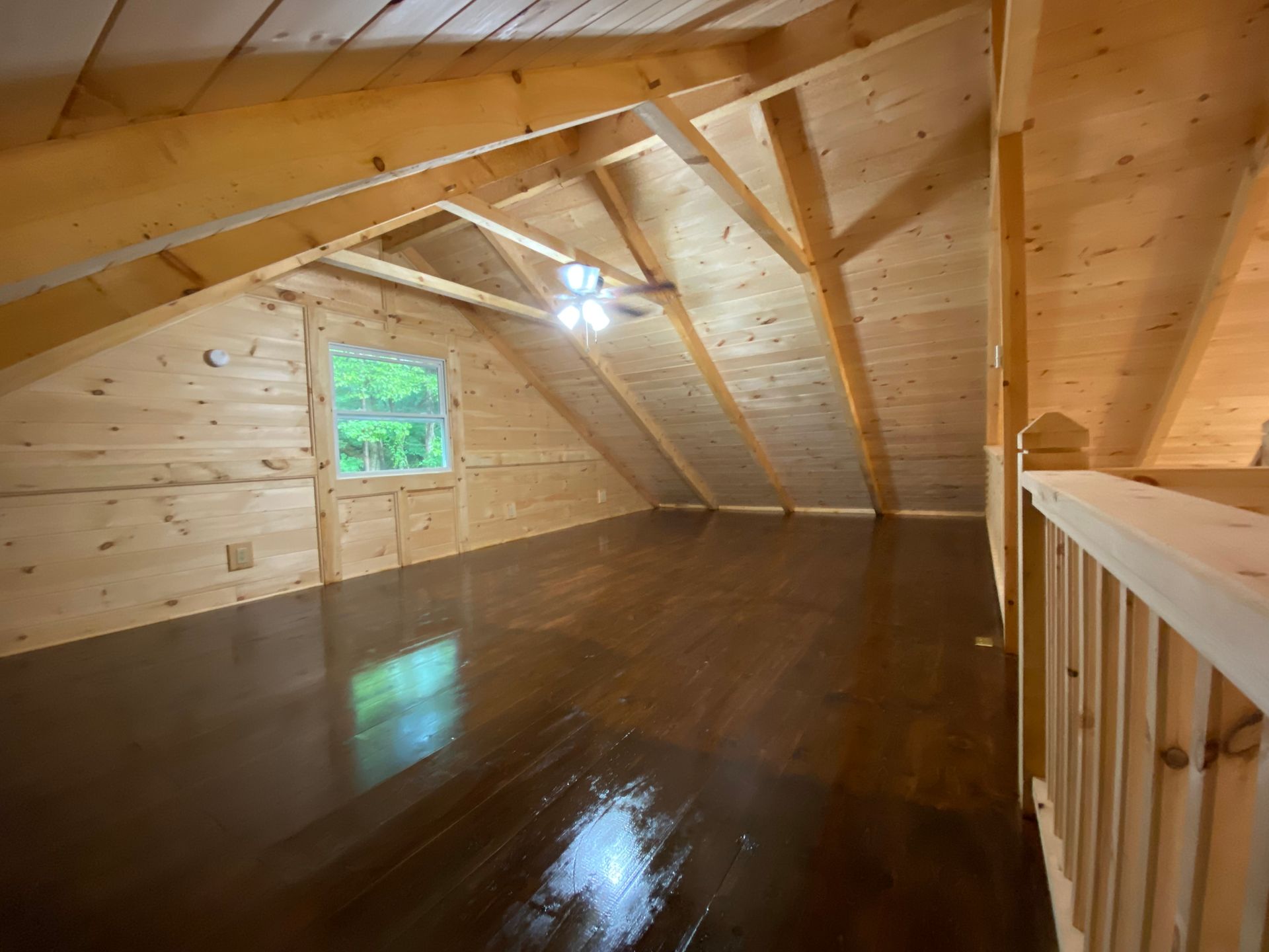 Interior of a wood-paneled cabin loft with stained wooden floor, a window, and a ceiling fan.