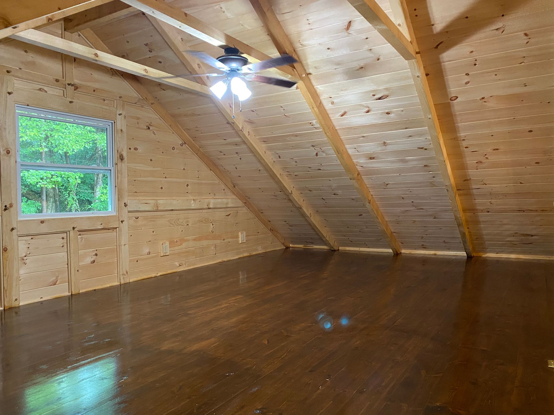 Wooden-paneled attic room with hardwood floor, window, and ceiling fan.