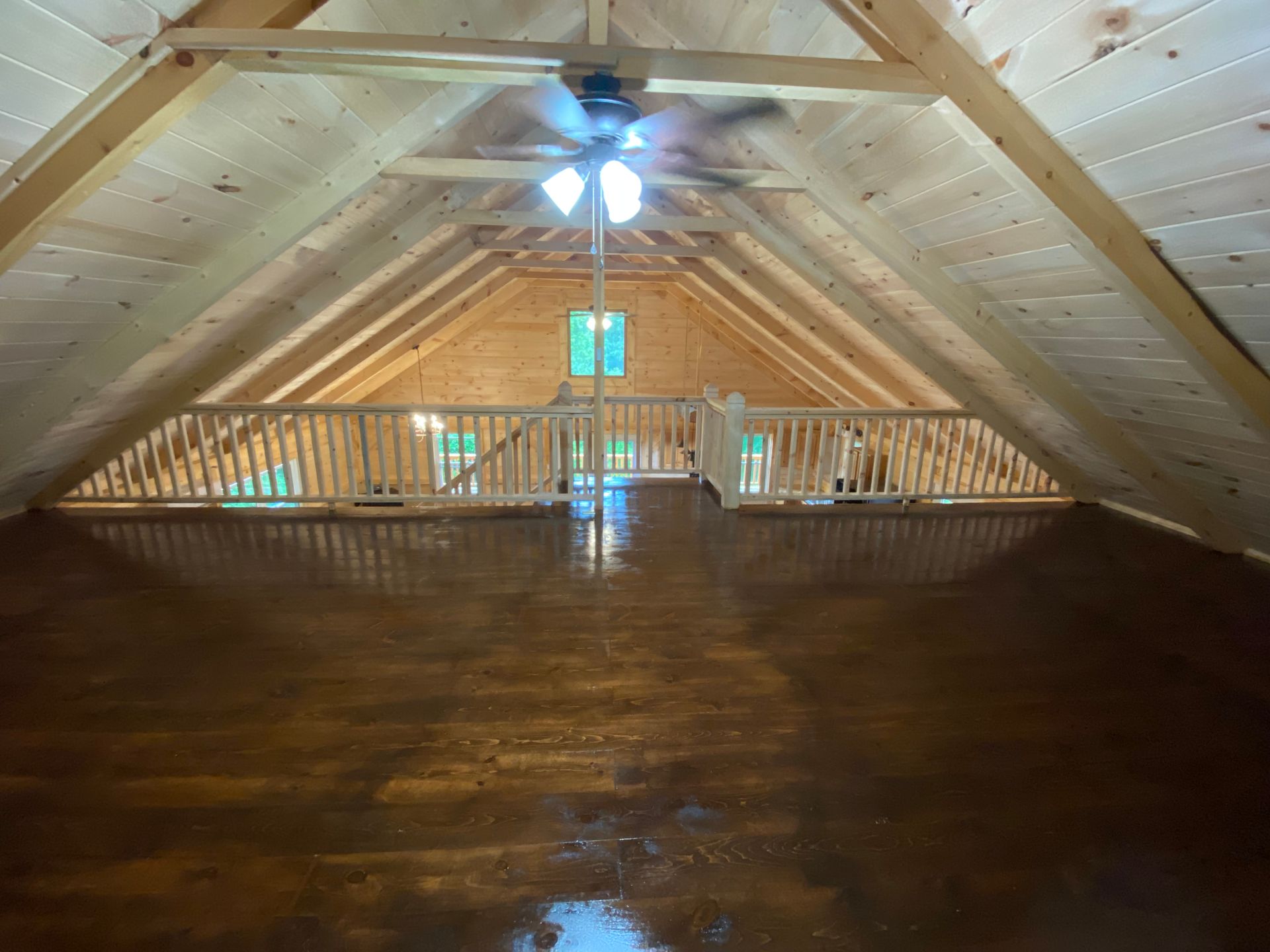 Wooden loft interior with glossy brown floor, exposed rafters, and a ceiling fan. Railing and window visible.