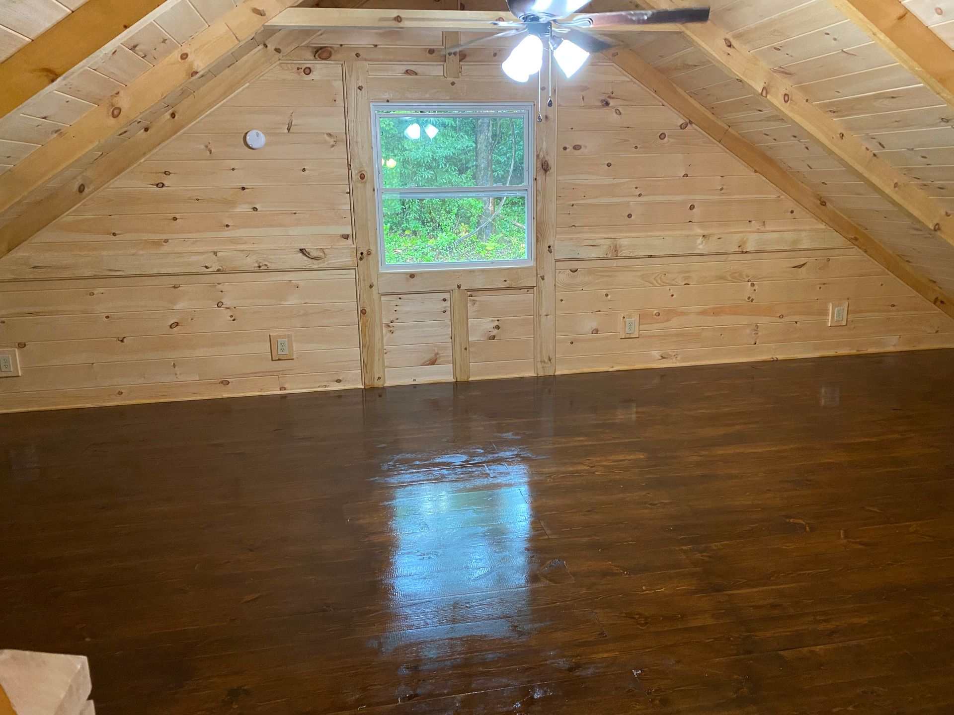 A-framed room with wood walls and dark floor, centered window, and ceiling fan.