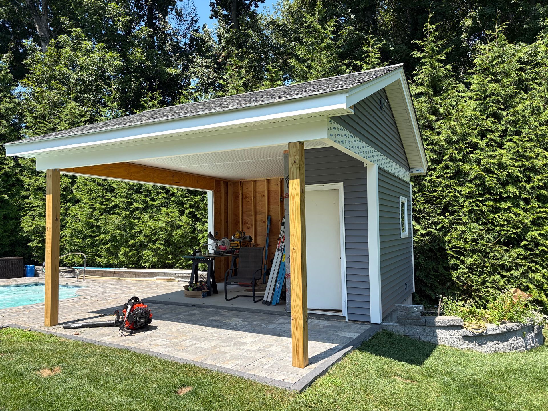 Small gray outbuilding with open front porch, white door, and tools on a concrete slab beside trees