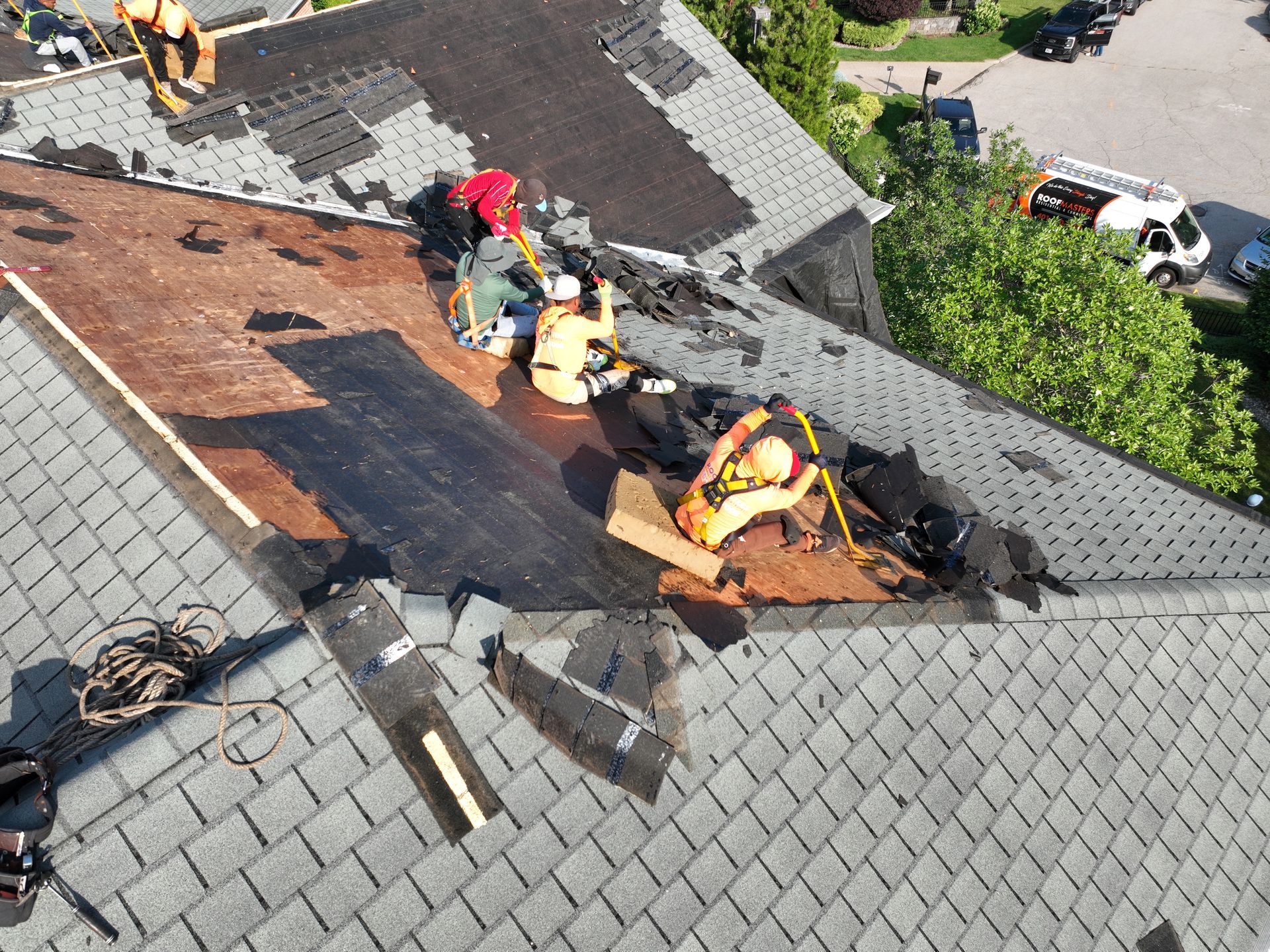 Roofers repairing a damaged shingle roof, with tools and tarps on a steep gray rooftop.