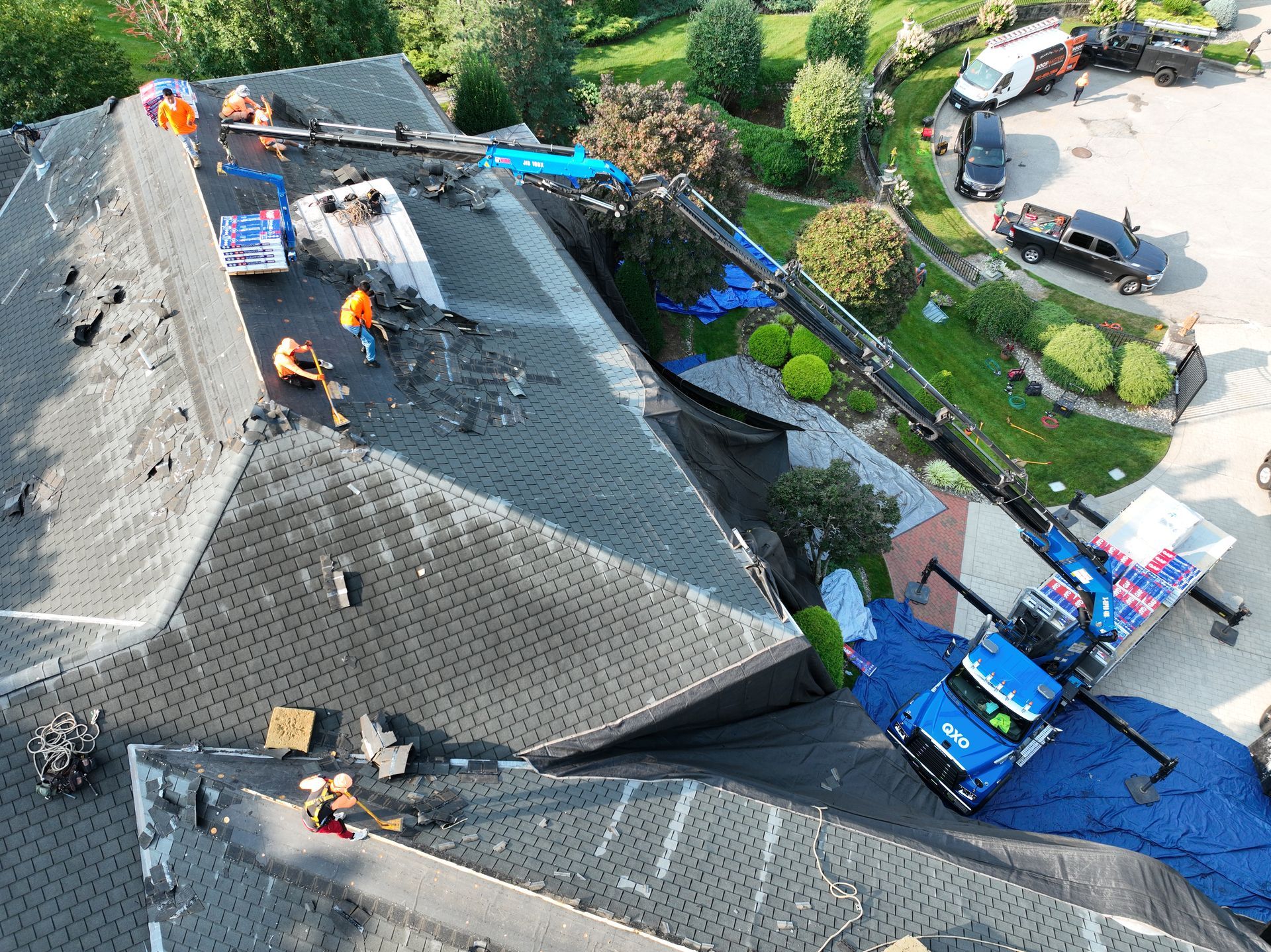 Roofers working on a steep gray shingle roof beside a blue tarp and driveway below