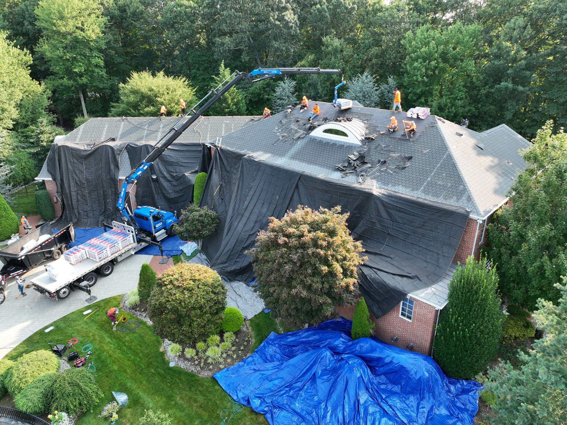 Workers tarp a large roof while a crane lifts materials beside a house in a wooded yard