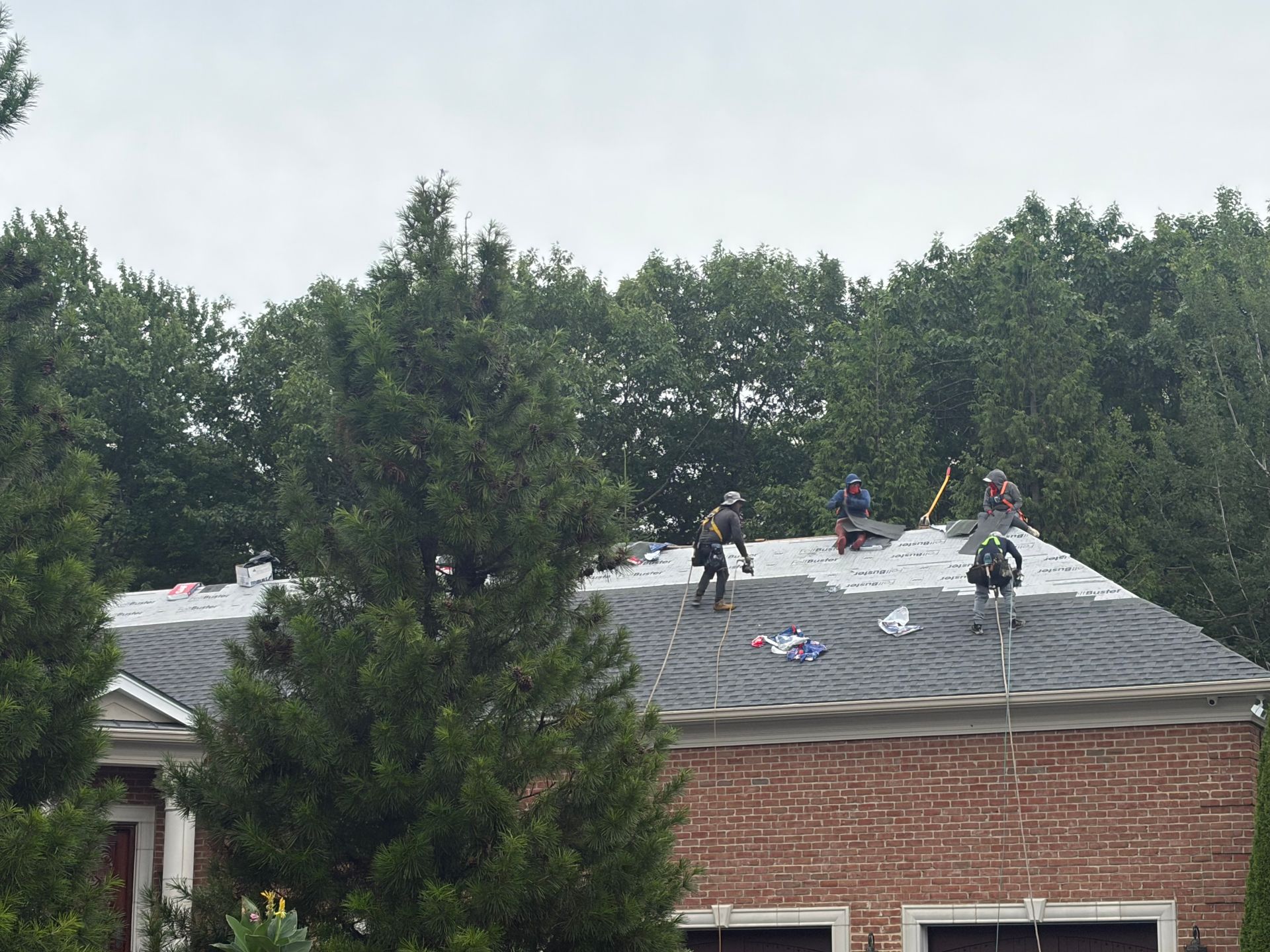 Three workers on a brick roof, repairing shingles near trees