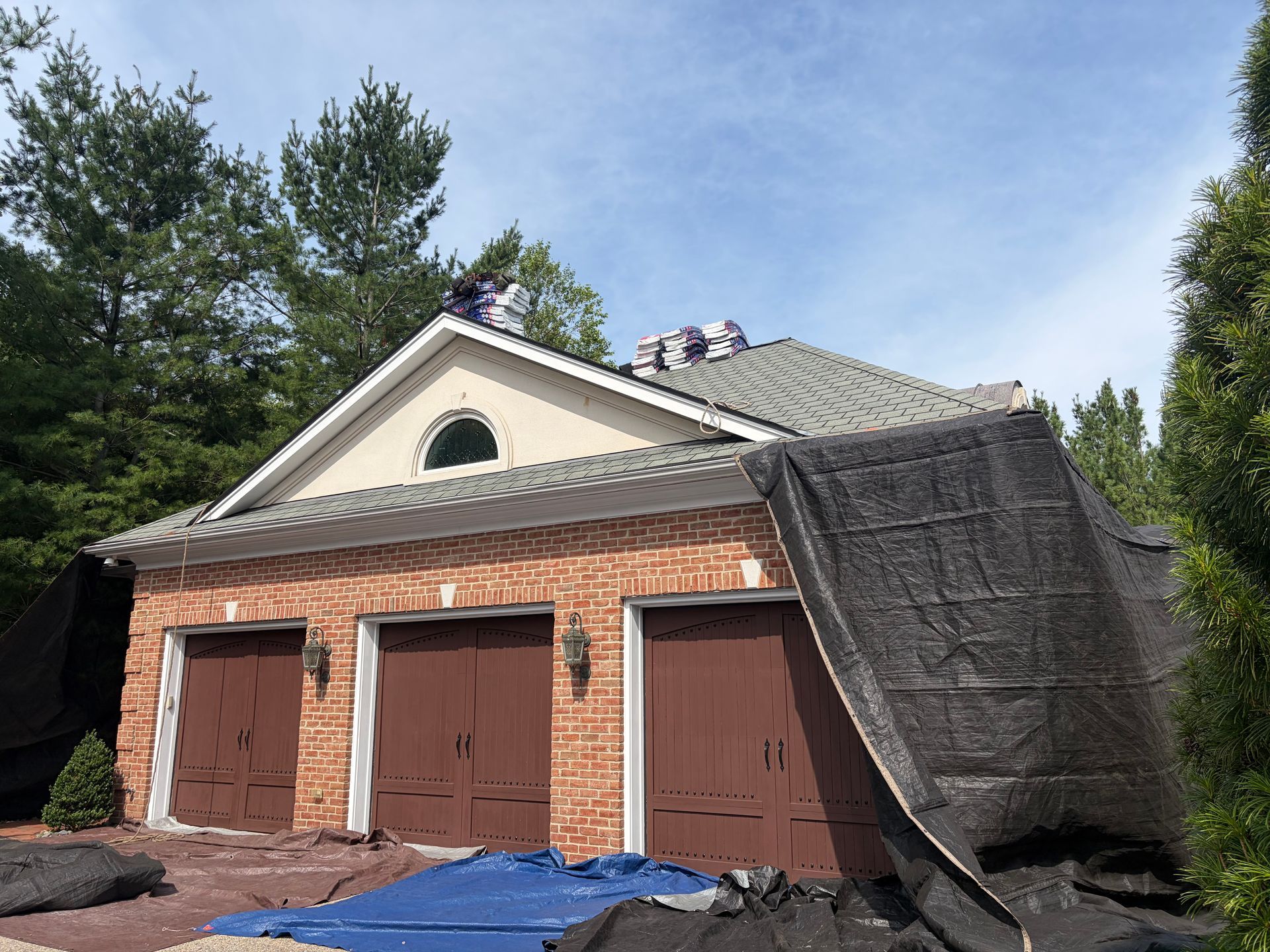 Brick garage with three brown doors and a steep, tarp-covered side roof among trees