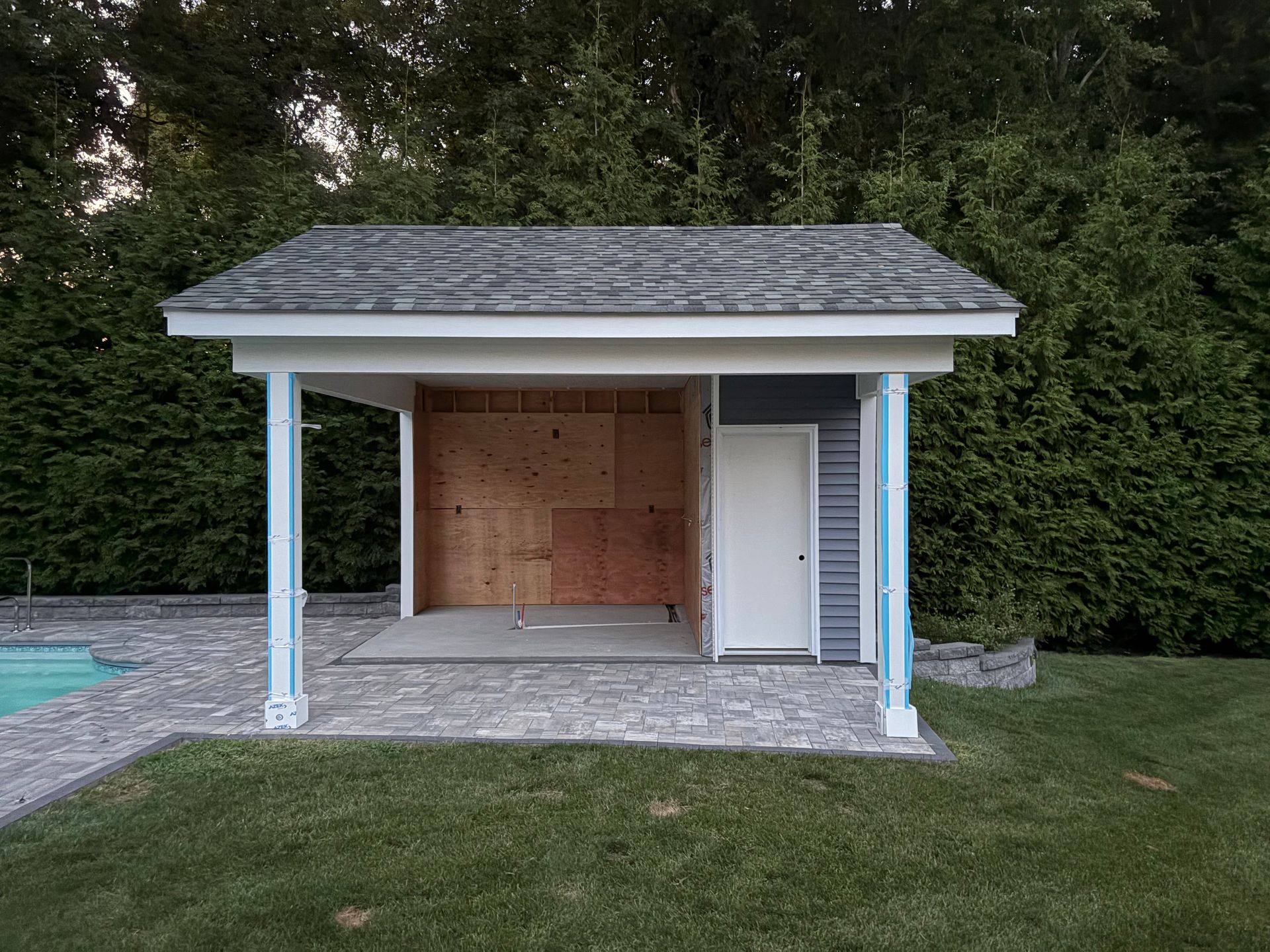 Small poolside pavilion with white posts, shingled roof, and enclosed storage against a wooded backdrop