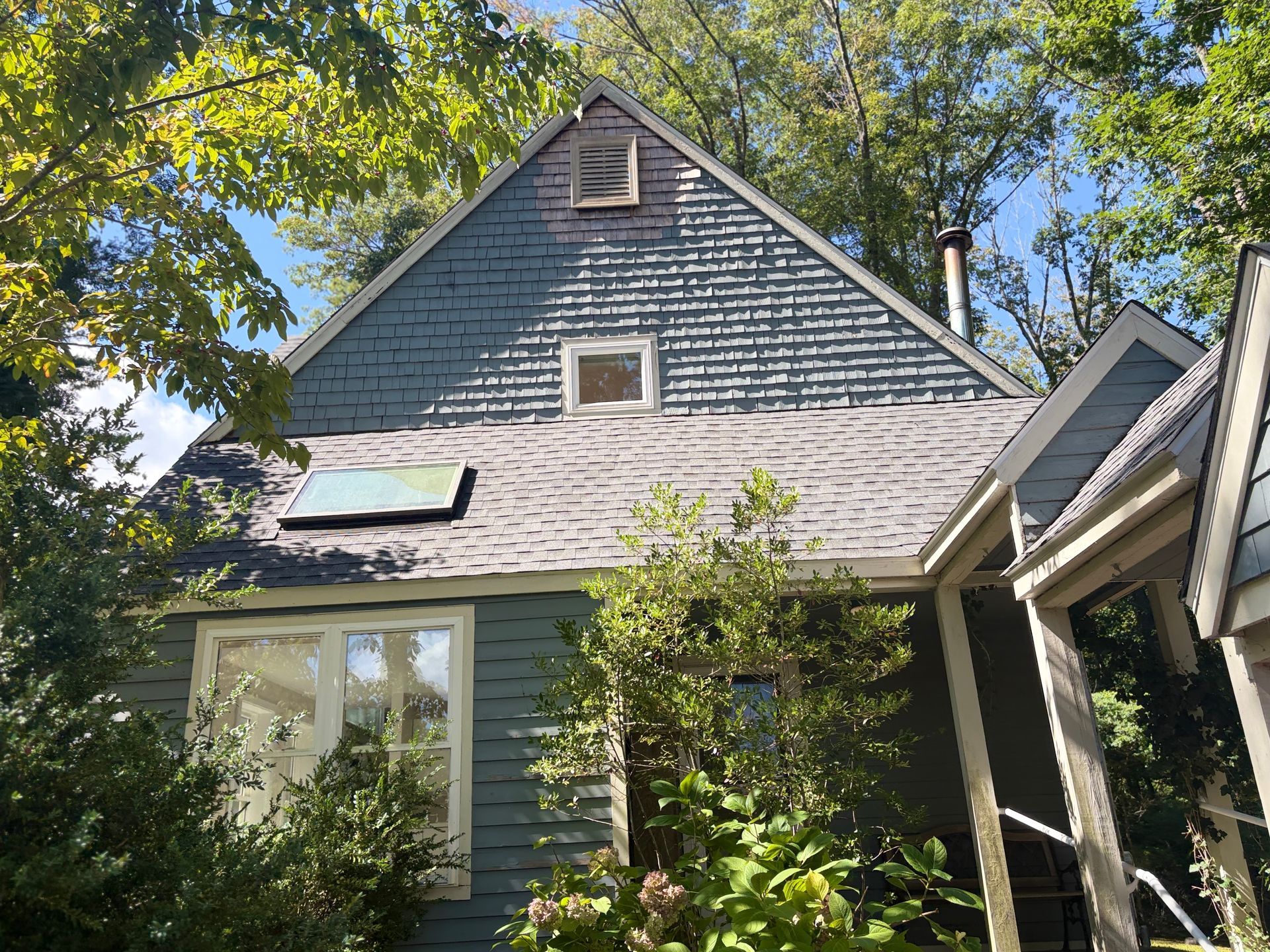 Small gray house with a steep roof and skylight, partly hidden by trees and greenery
