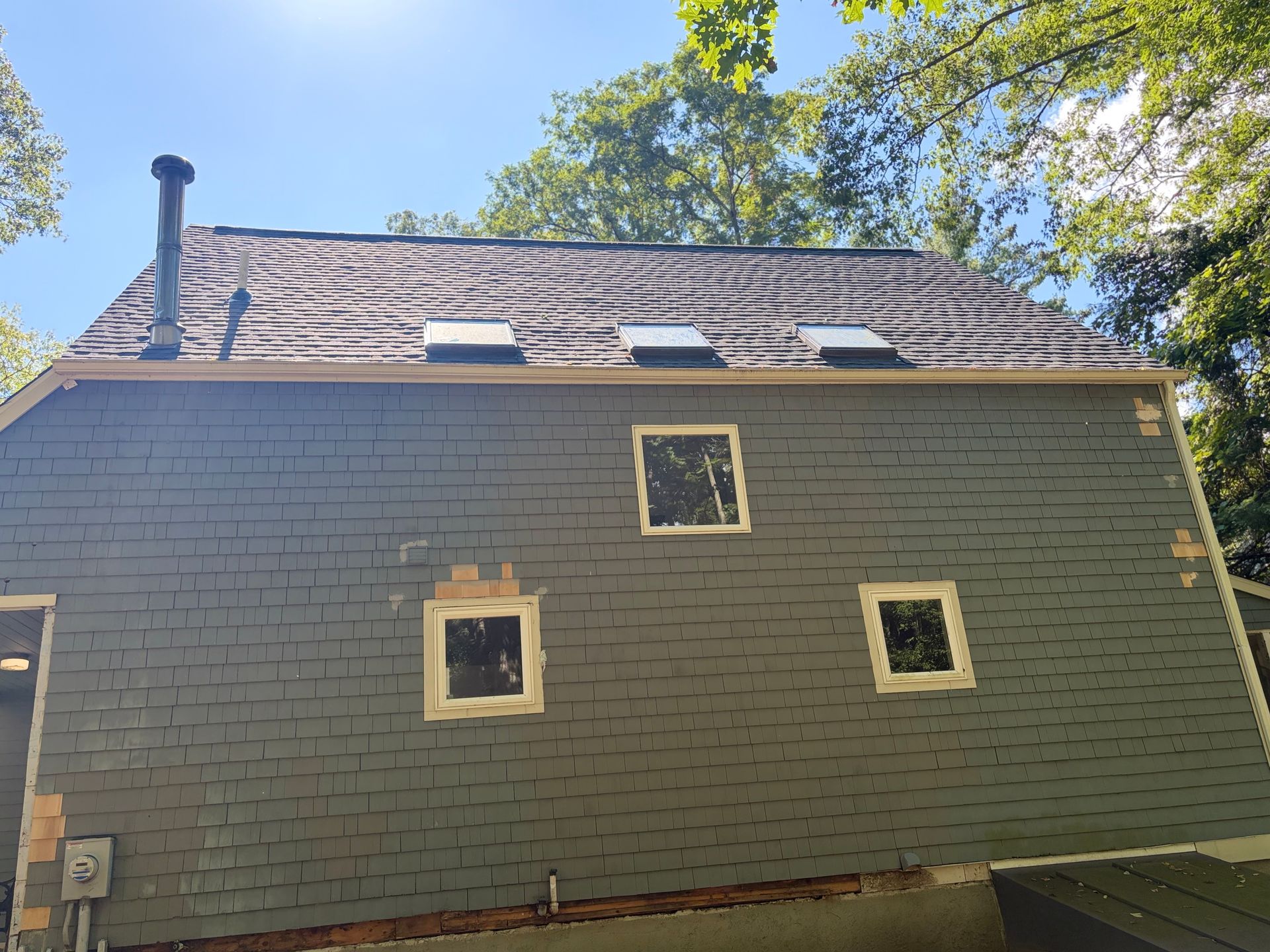 Back of a gray house with small windows and a shingled roof under trees