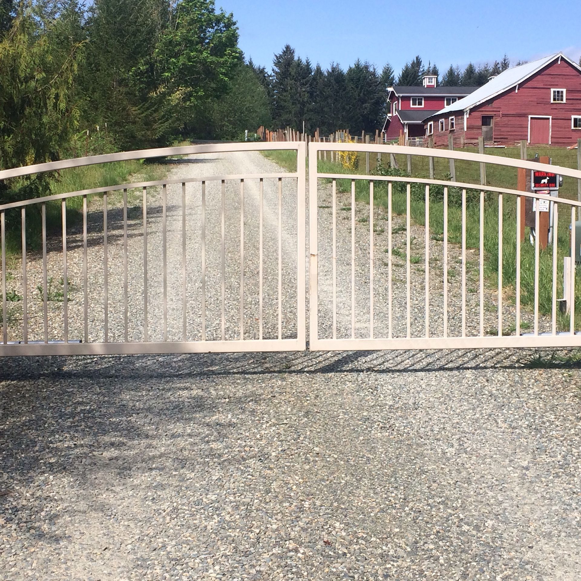 A white gate is open to a gravel road with a red barn in the background